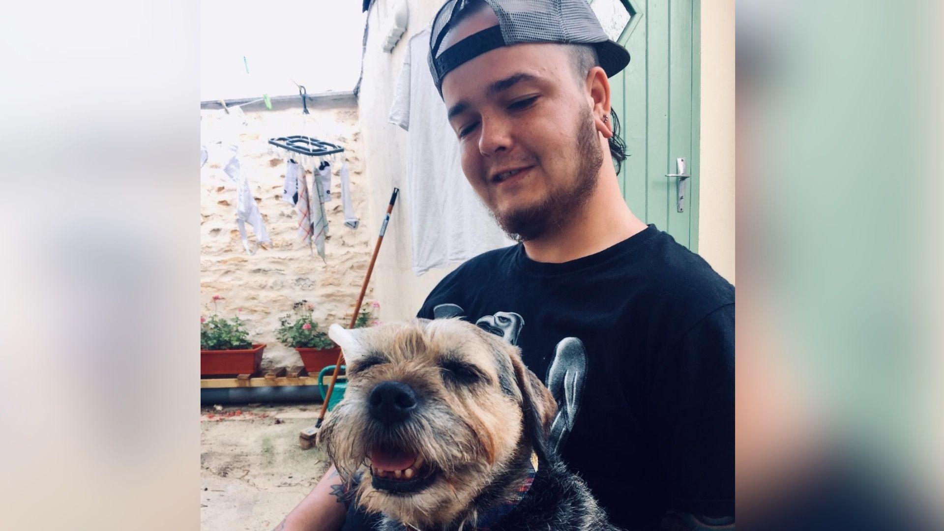 Hobie Harrison sits in front of his parents yard with plants in potted containers behind him. He is wearing a baseball cap the wrong way around and has a black T-shirt on. He is looking at a little terrier type dog which has tan and black colouration