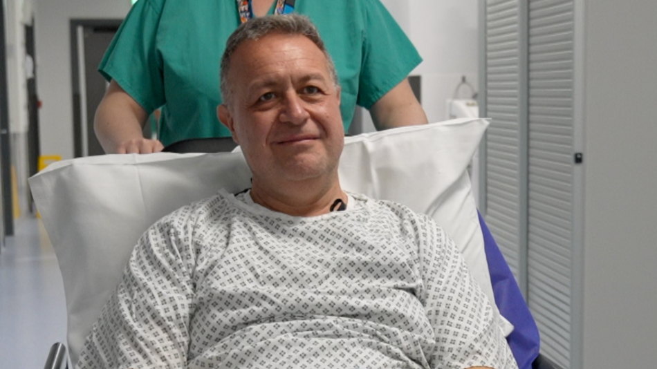 Steve wears a hospital gown and sits up on a gurney. He is propped up on a pillow and is wearing a patterned hospital gown in black and white. He is being pushed on his way to have his operation