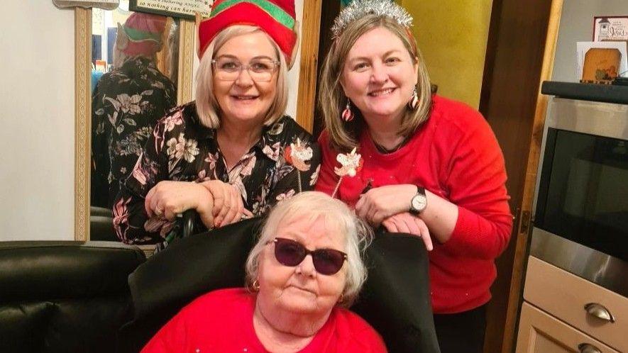 Two women - Brendya young and her sister Janice - wearing Christmas hats, behind their mother Margaret Chisholm who also has Christmas-related items on her head, and dark glasses.