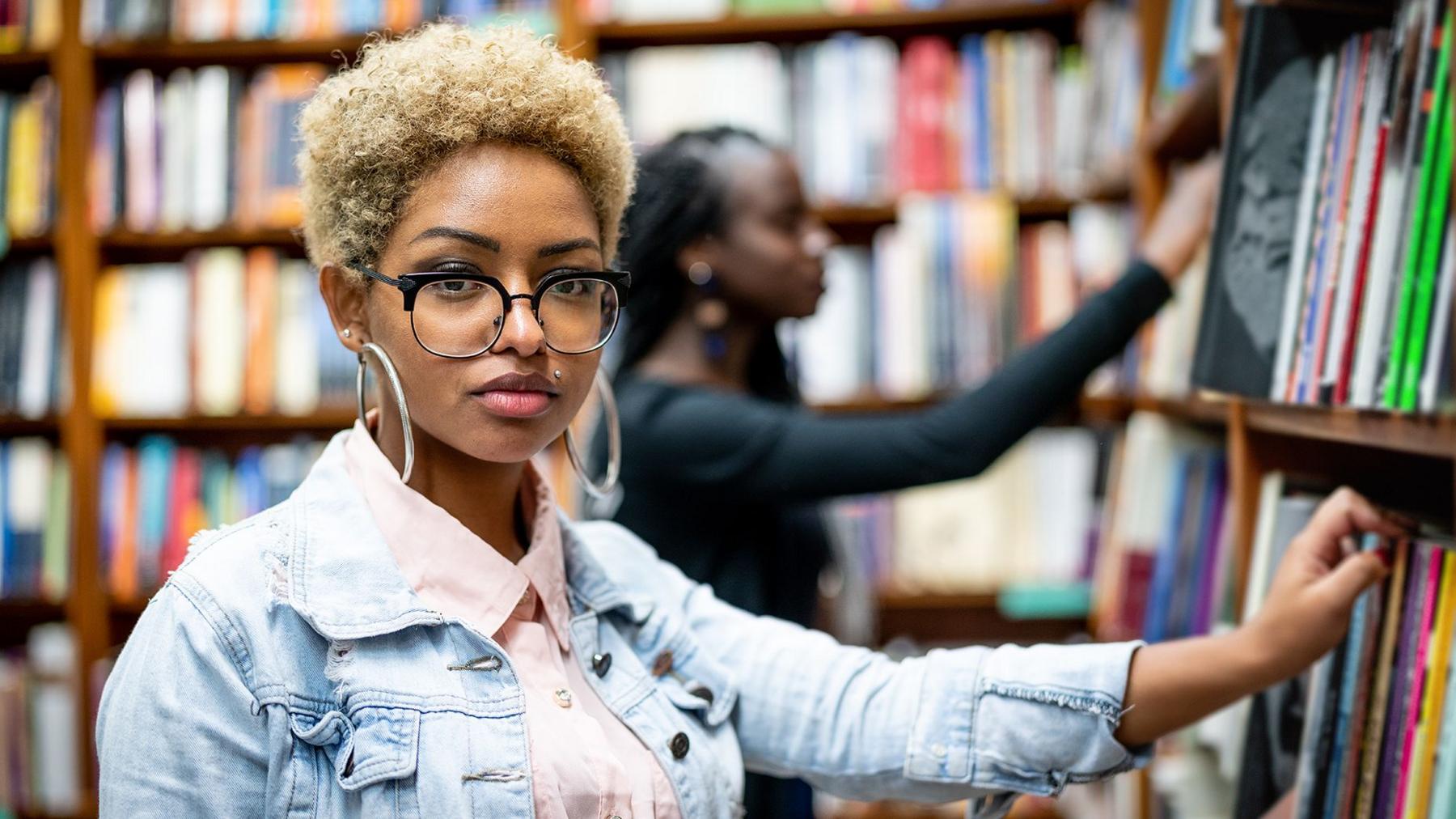 A female university student in a library wearing a demin jacket looking towards the camera with her hand on a book