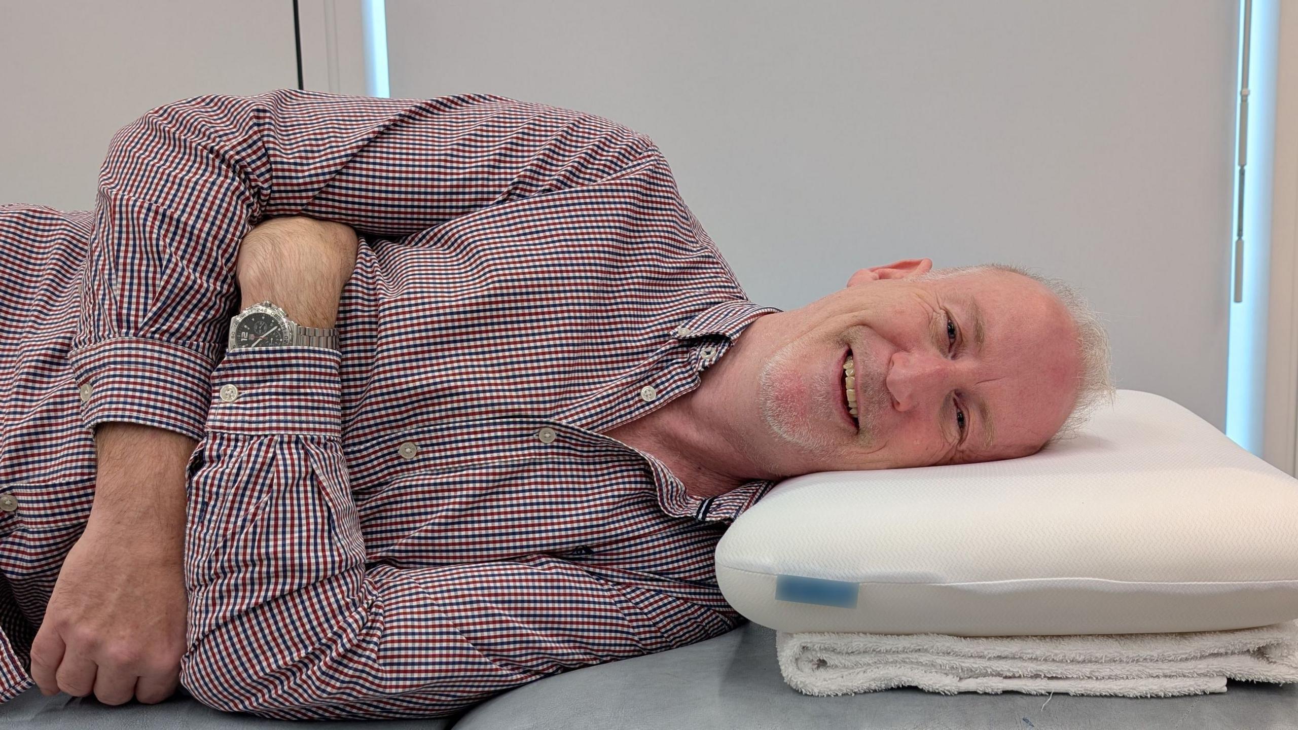 Person lying on their side on a padded treatment table with their head resting on a foam pillow. The person is wearing a checked button-up shirt and a wristwatch, with arms folded across the torso. The pillow is positioned on a folded towel. The window blinds in the background are drawn.