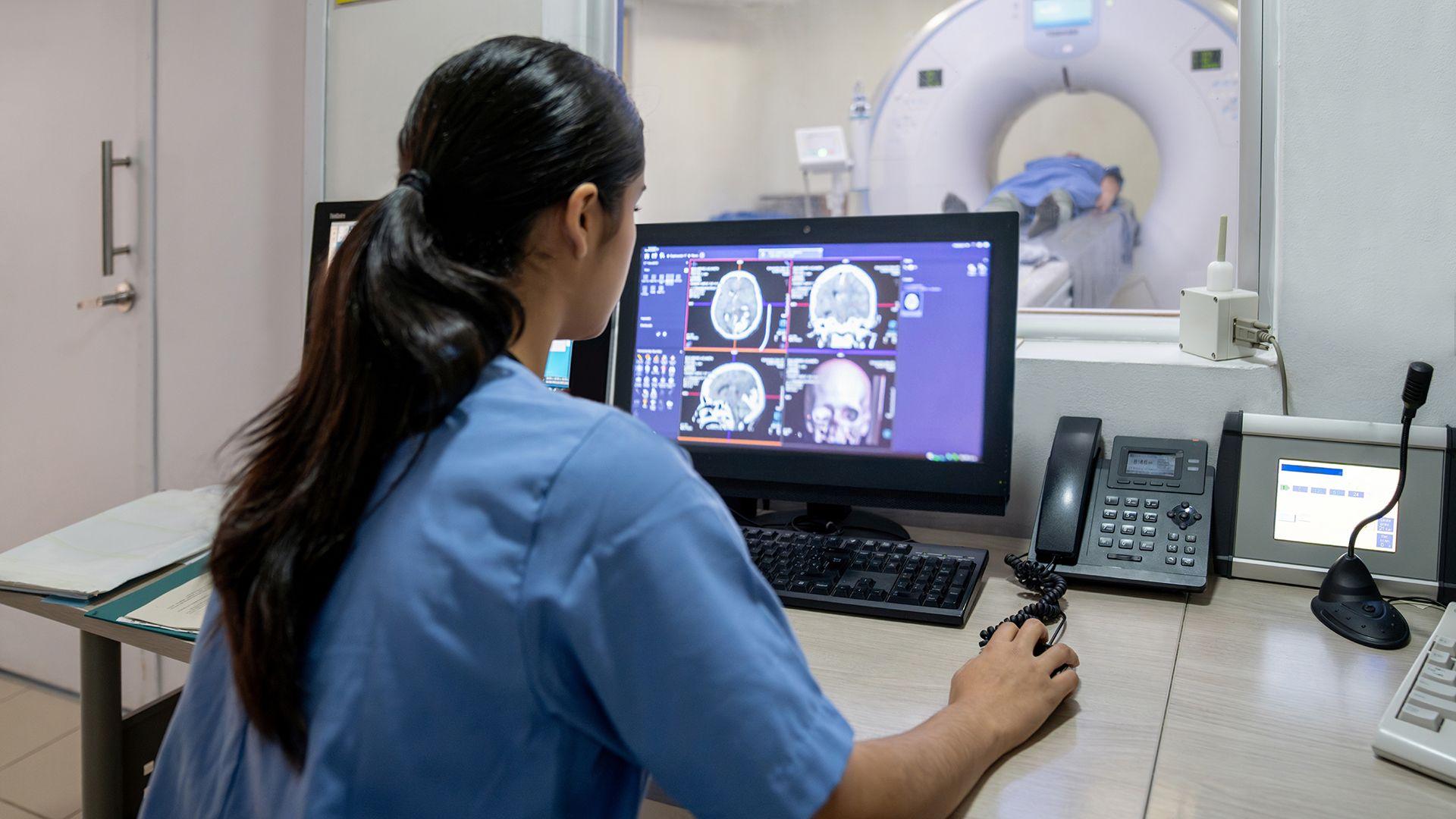 Stock photo shows a person in a doctor's overall using a computer that shows brain scans, with a person being inserted into an MRI machine in the background in a hospital environment.