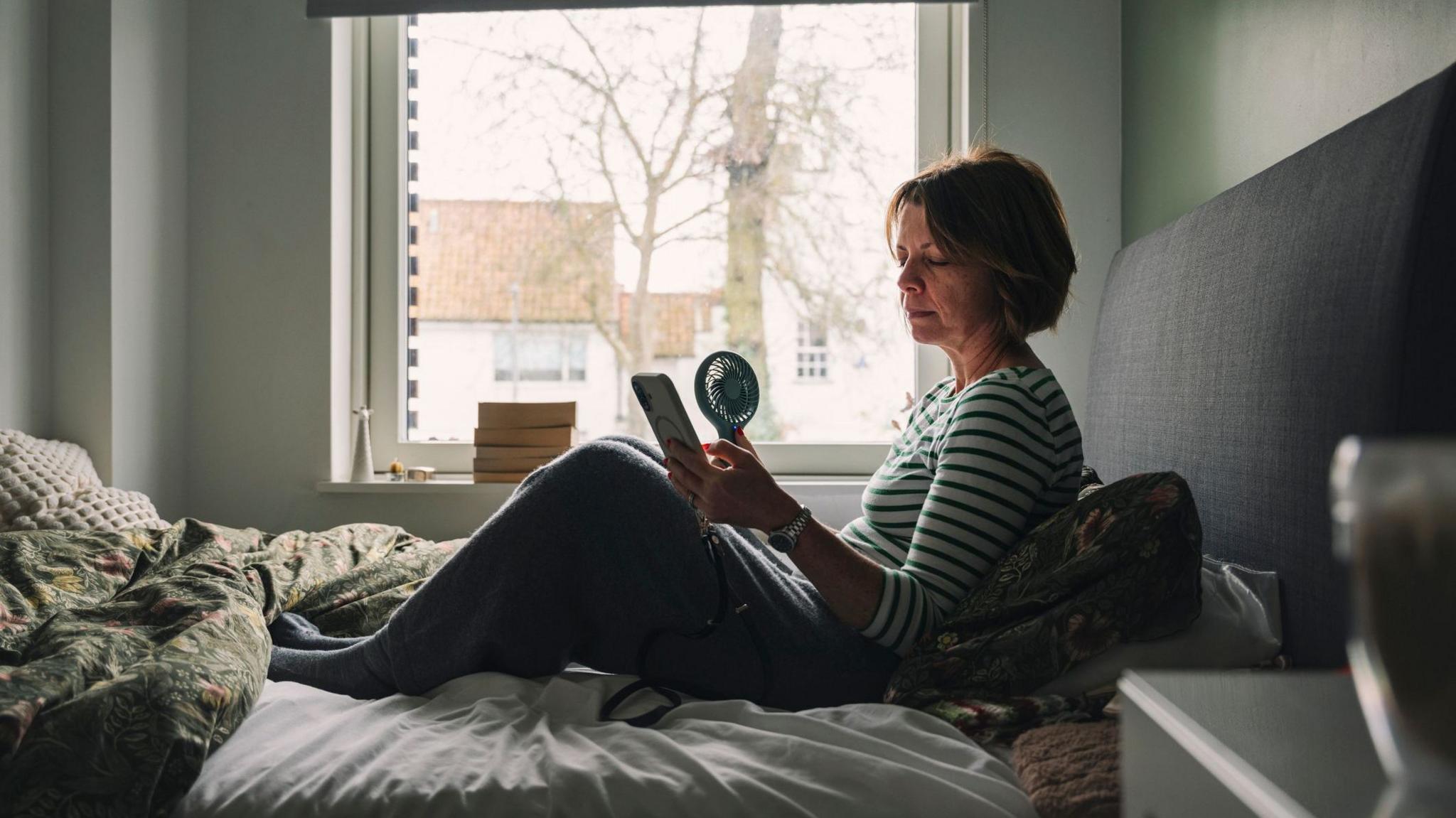 A stock photo of a woman in bed with a handheld electric fan.