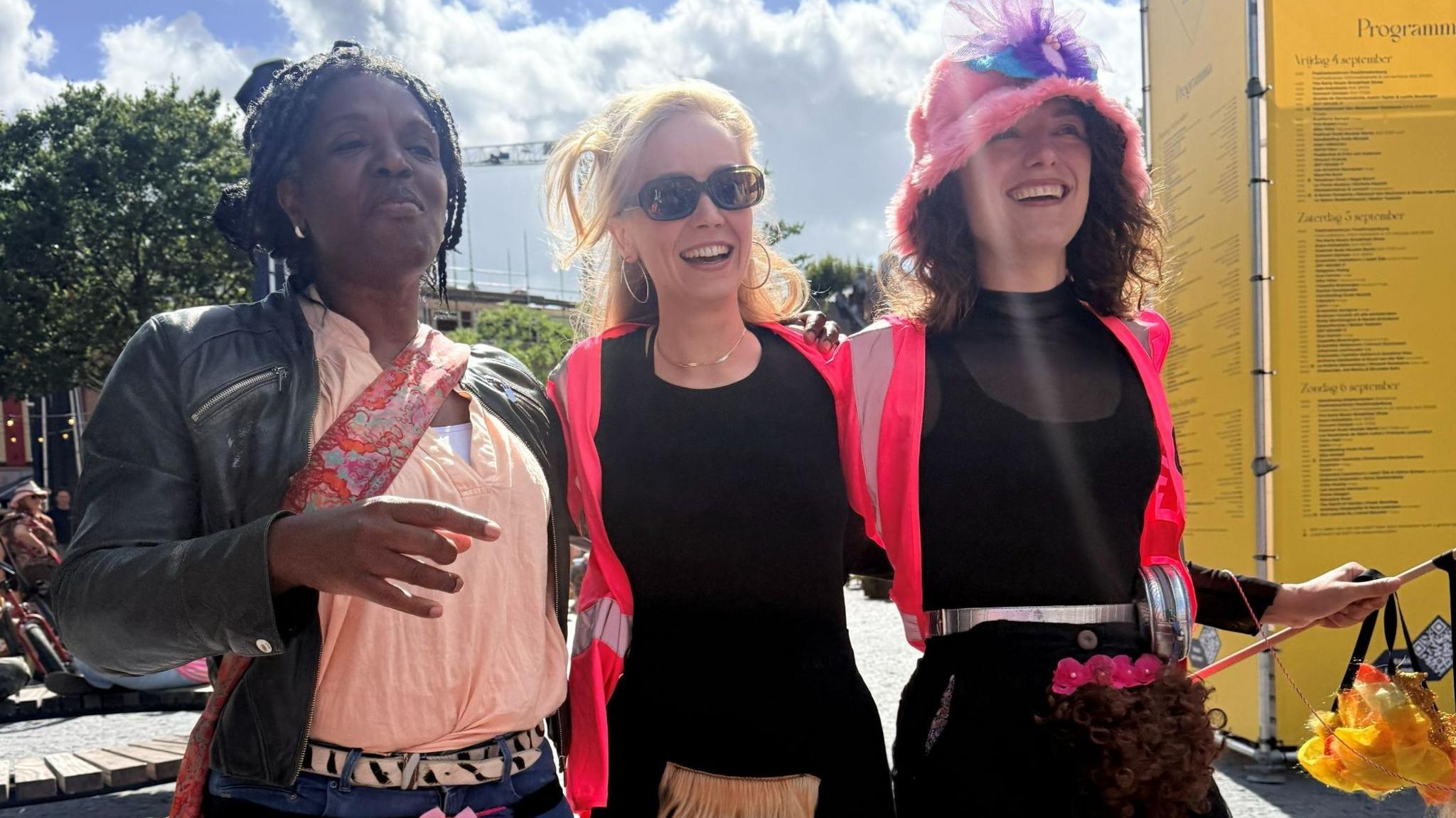 Three people who joined in the march. Two wear bright pink jackets, the other a pale pink shirt. They are smiling and wearing merkins - artificial pubic hair pieces - which sits over their clothing.
