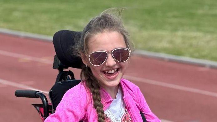 Tammy is sat in a black wheelchair on an athletics track which is red. She has long brown hair in a plait, and is wearing a white top with a pink butterfly on and a pink jacket. She is looking at towards the viewer smiling, and is wearing sunglasses.