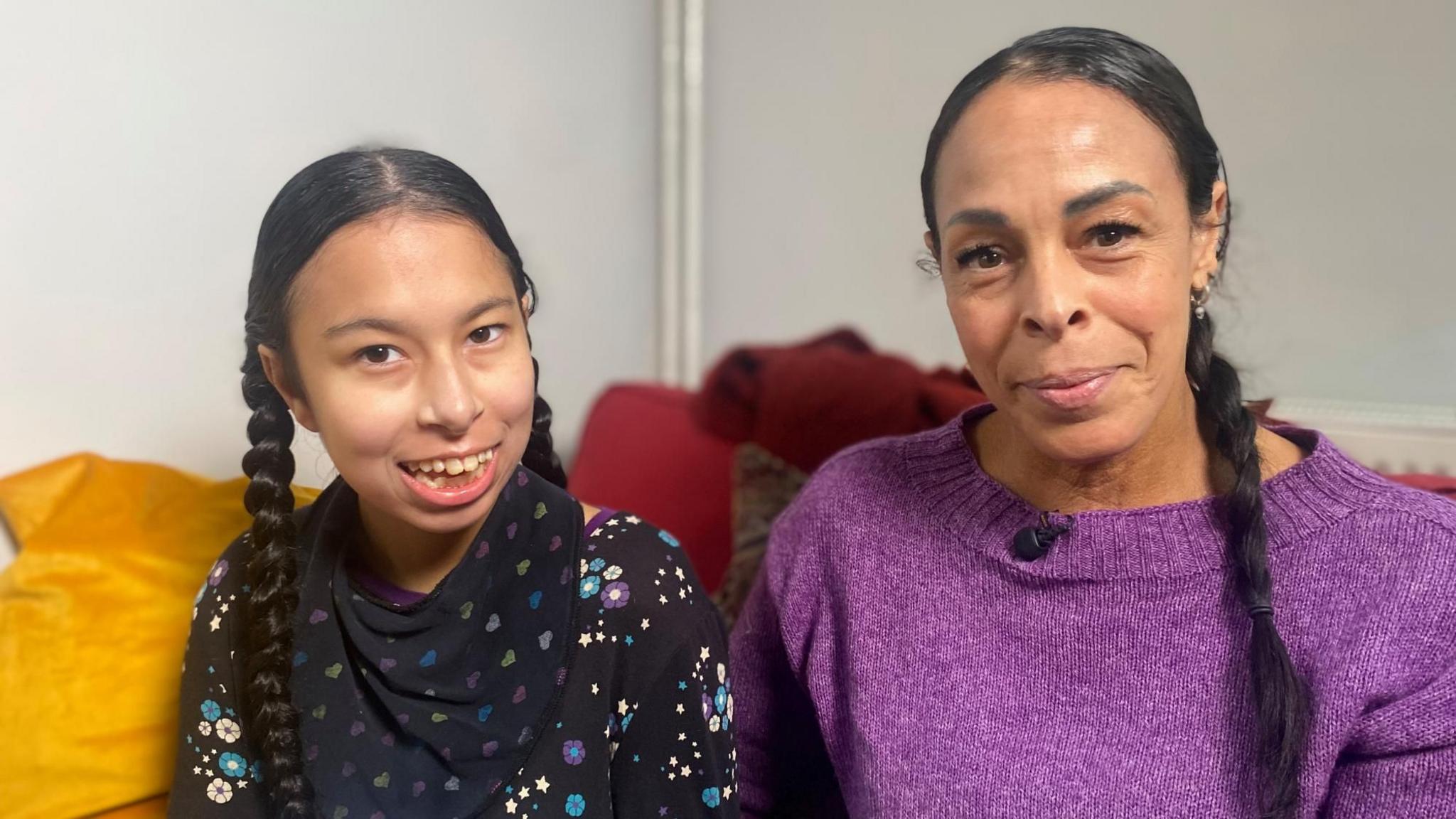 Neave and Natasha are sitting indoors on a sofa. Both have long, dark hair styled in braids/pigtails. Natasha, who is taller, is wearing a purple knitted top with a small microphone clipped near the neckline. Neave is dressed in a black top patterned with colourful stars and flowers. They are both looking at the camera and smiling.