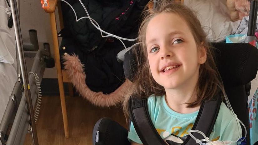 Tammy is sat in a black wheelchair looking at towards the viewer smiling. She has long brown hair, and is wearing a light blue top. Behind her is a hospital bed on the left.