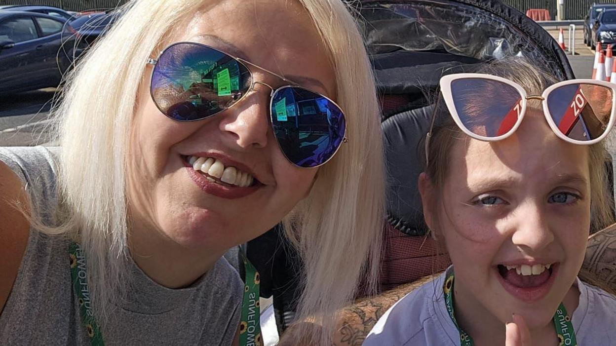 On the left is Lynn, Tammy's mother, next to her daughter. They are standing in a car park taking a selfie. Lynn has shades on, Tammy also has shades but they are placed on the top of her head.