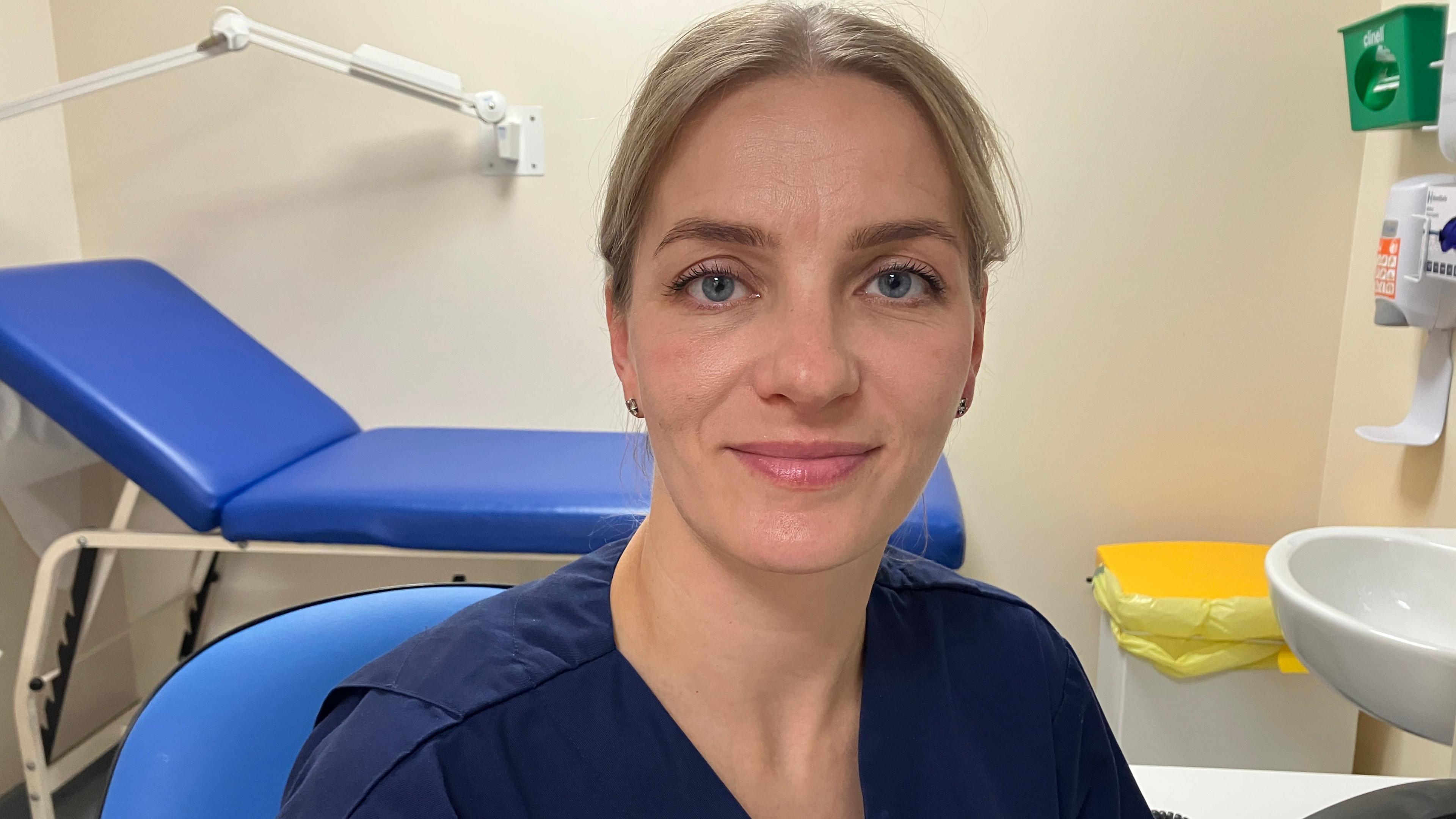 A midwife sat inter office wearing blue scrubs. She has blonde hair tied back and is smiling at the camera