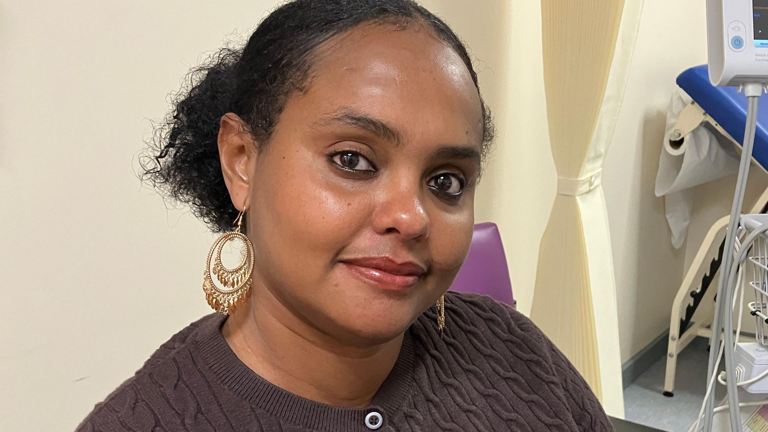 A woman with black hair, tied back, large gold earrings is smiling at the camera. She is wearing a brown cardigan.