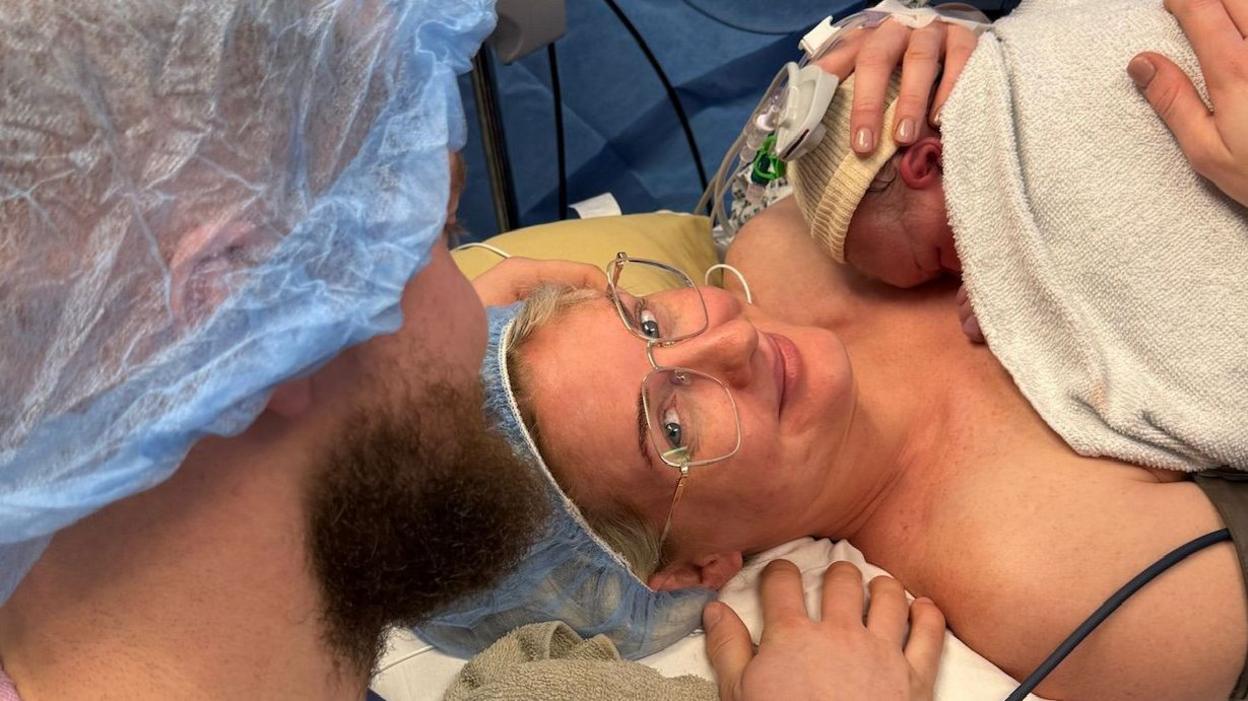 A photo of woman holding her new born baby after giving birth, with the back of her partners head in scrubs.