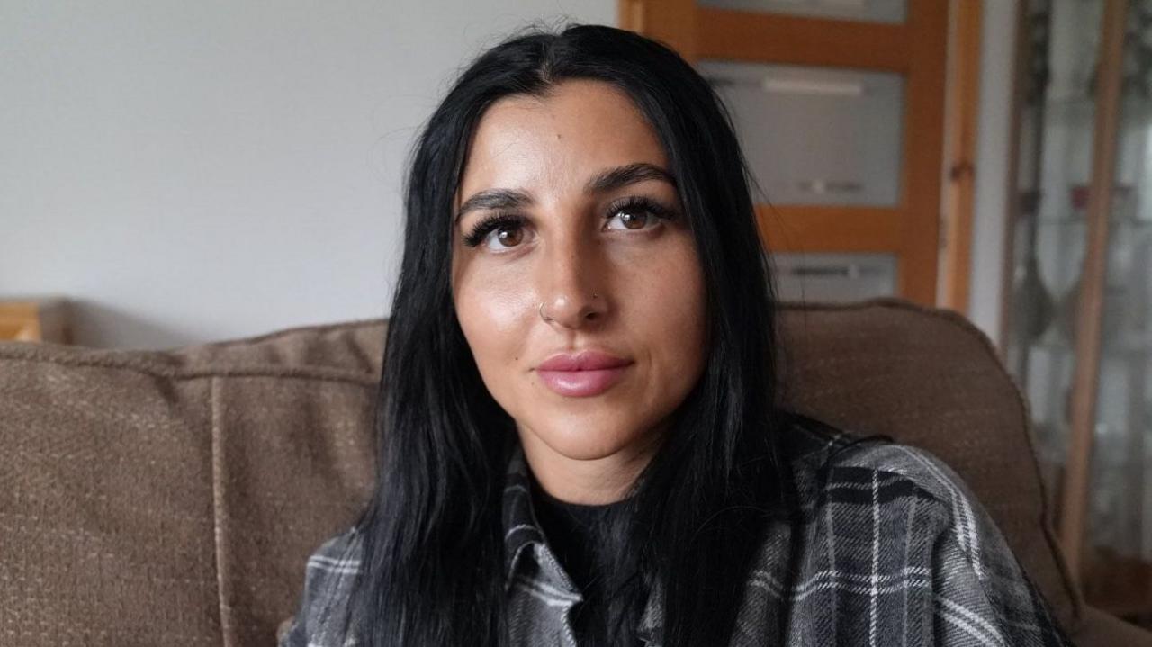 A woman with long black hair wearing a tartan shirt, sitting on the sofa.