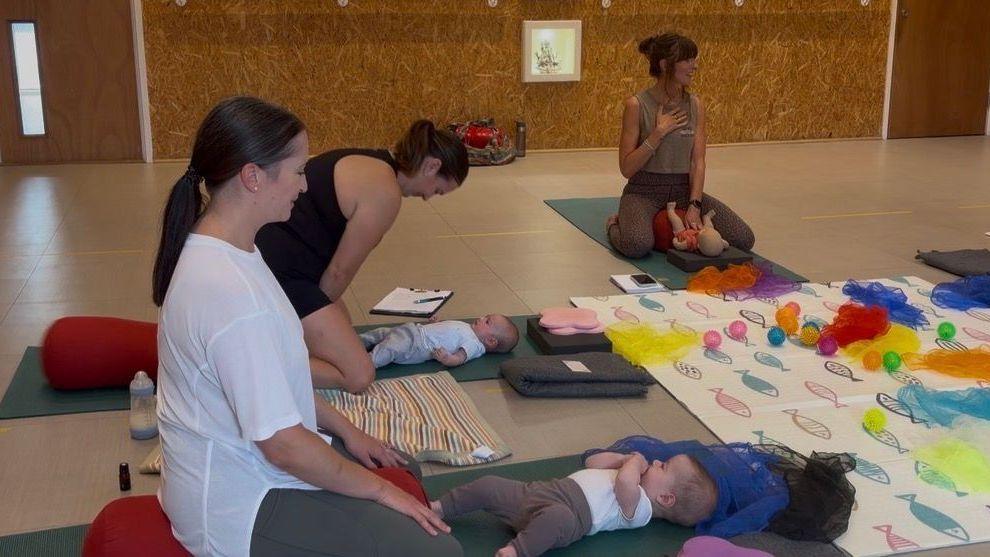 Mother and baby yoga with an instructor sitting in front of the class and two mothers and their babies following instructions.