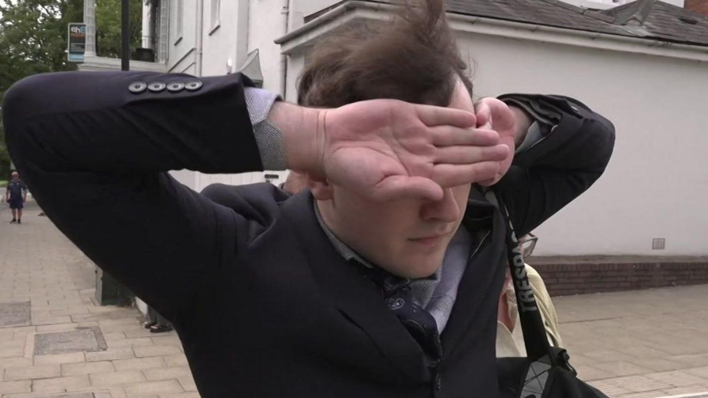 A white man with brown hair with a dark suit and blue shirt puts his hands over his eyes as he walks down the street 