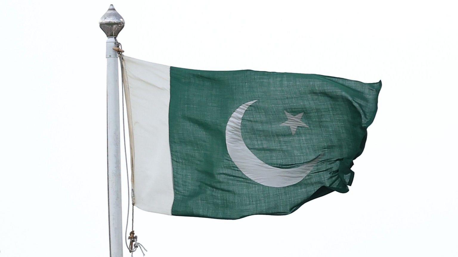 A Pakistan flag flying on a flag-pole. It is green with a white border down the left hand side, with a crescent moon and a five-pointed star in its centre.
