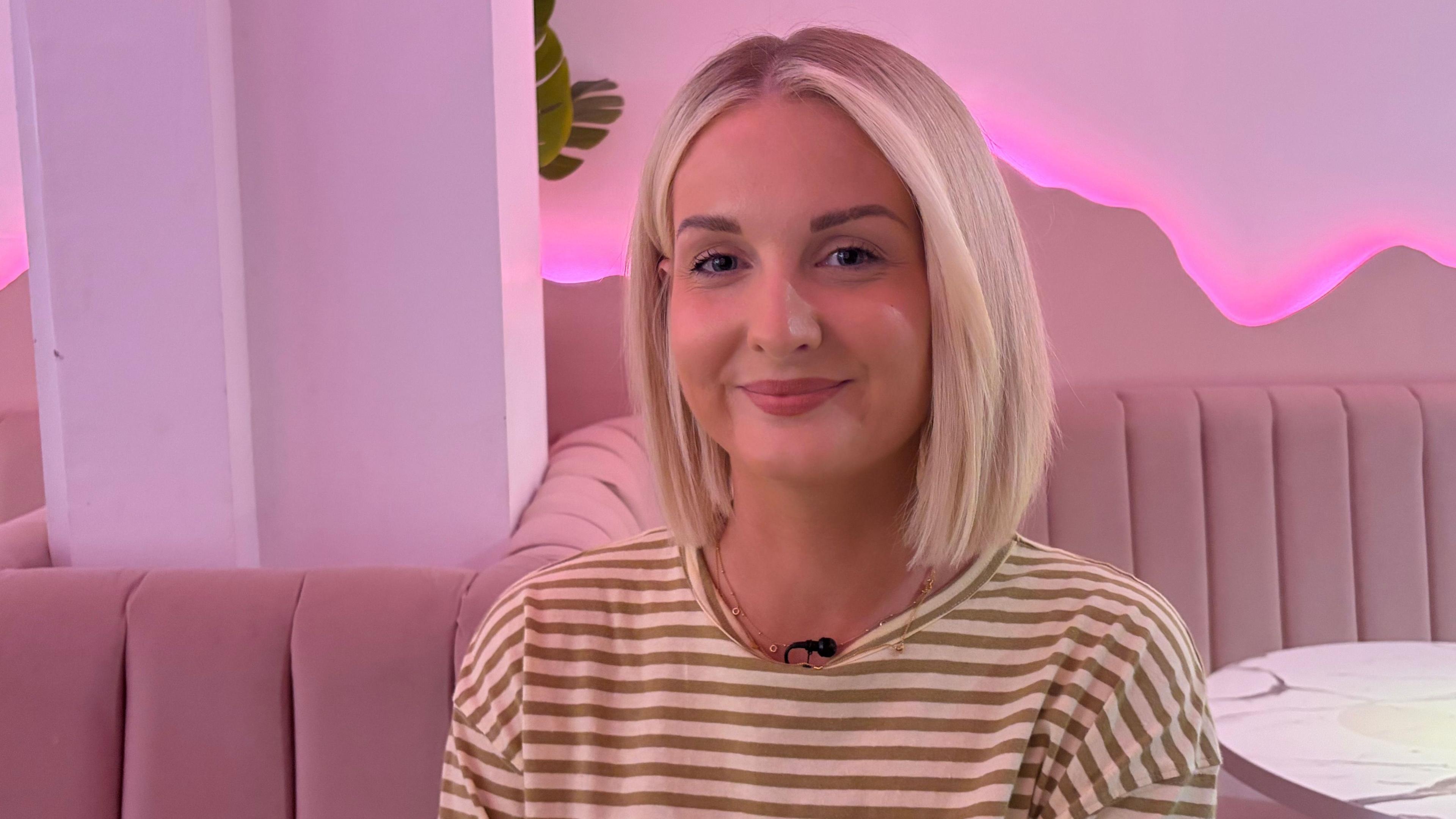 A picture of a woman with short blonde hair in a stripy top sitting in front of a pink background