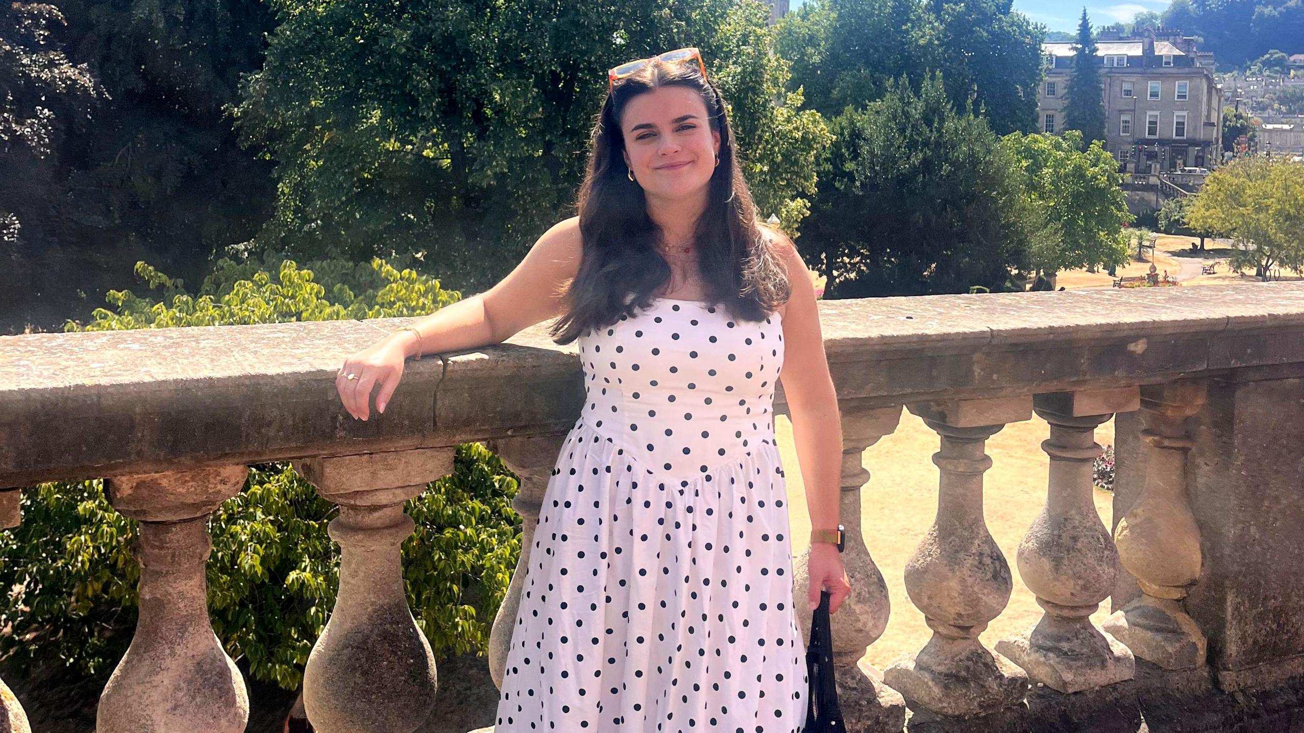 Ella Kipling, a young woman with long dark hair, smiles at the camera. She is wearing a long, white dress with black polkadots and is standing in front of a large park with old buildings in the background.