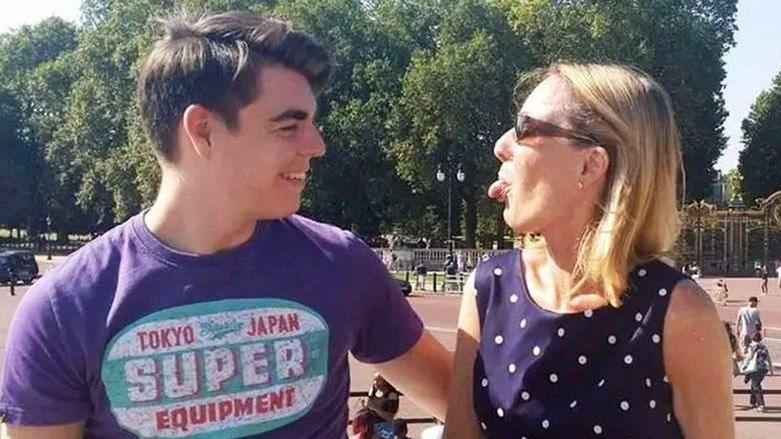 Oliver McGowan, wearing a purple t-shirt with a logo, is standing outside Buckingham Palace. He is staring at his mother Paula McGowan who is standing next to him and wearing a dark blue polka dot dress and sunglasses. She is playfully sticking her tongue out at her son and he is laughing.