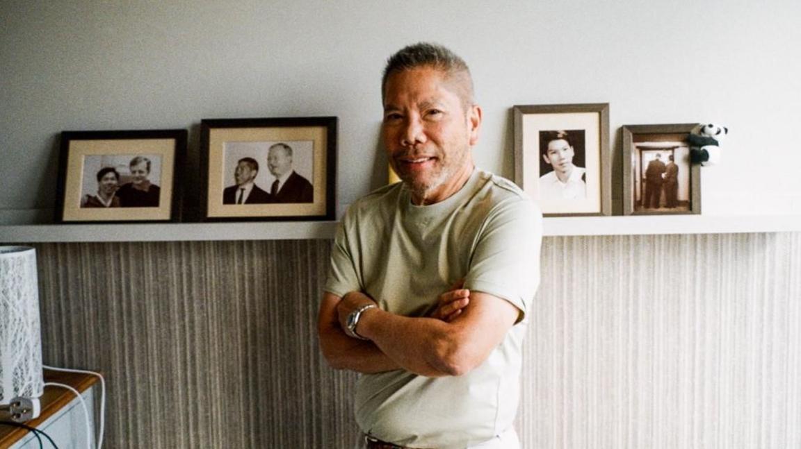 A man in his 70s crosses his arms and smiles at the camera. There is a picture shelf behind him featuring pictures of him and his late husband, as well as a portrait of the man in his younger years.