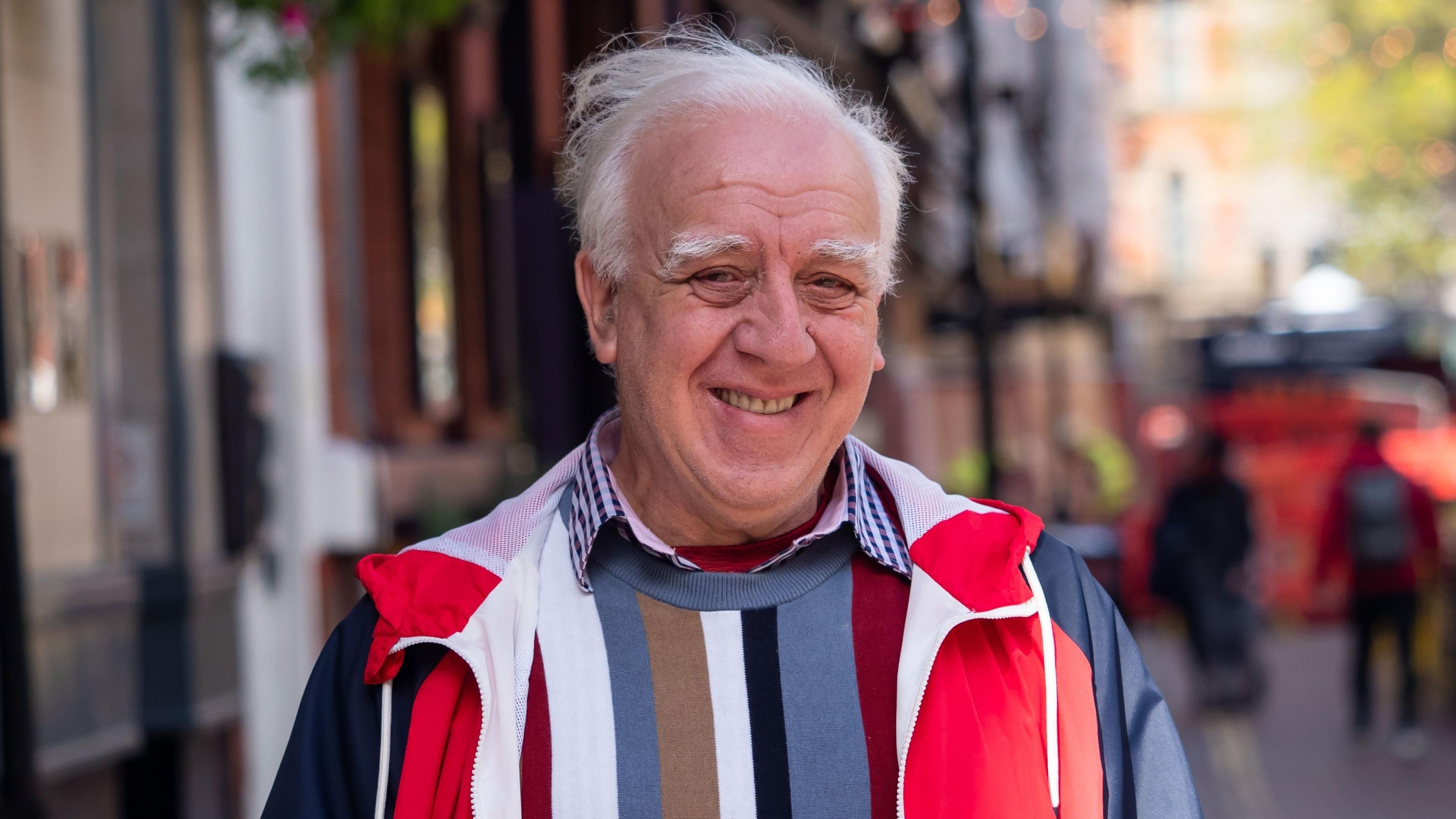 A man in his 70s smiles at the camera, the background behind him although blurred is Canal Street in Manchester's gay village. He's wearing a red and blue anorak, a checkered shirt and a striped jumper. He looks relaxed and happy.