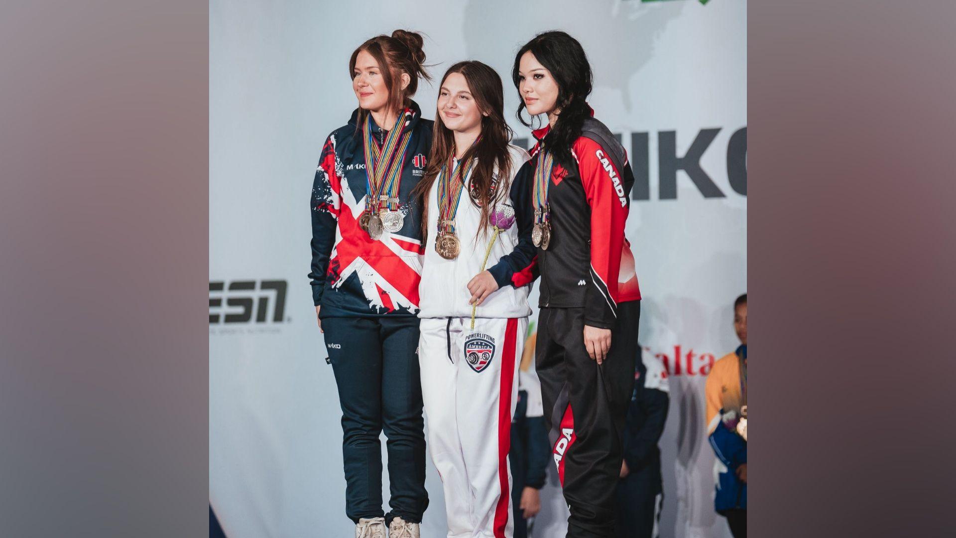 Three women stand on a podium in front of a banner for the International Powerlifting Federation. They all have medals around their necks.