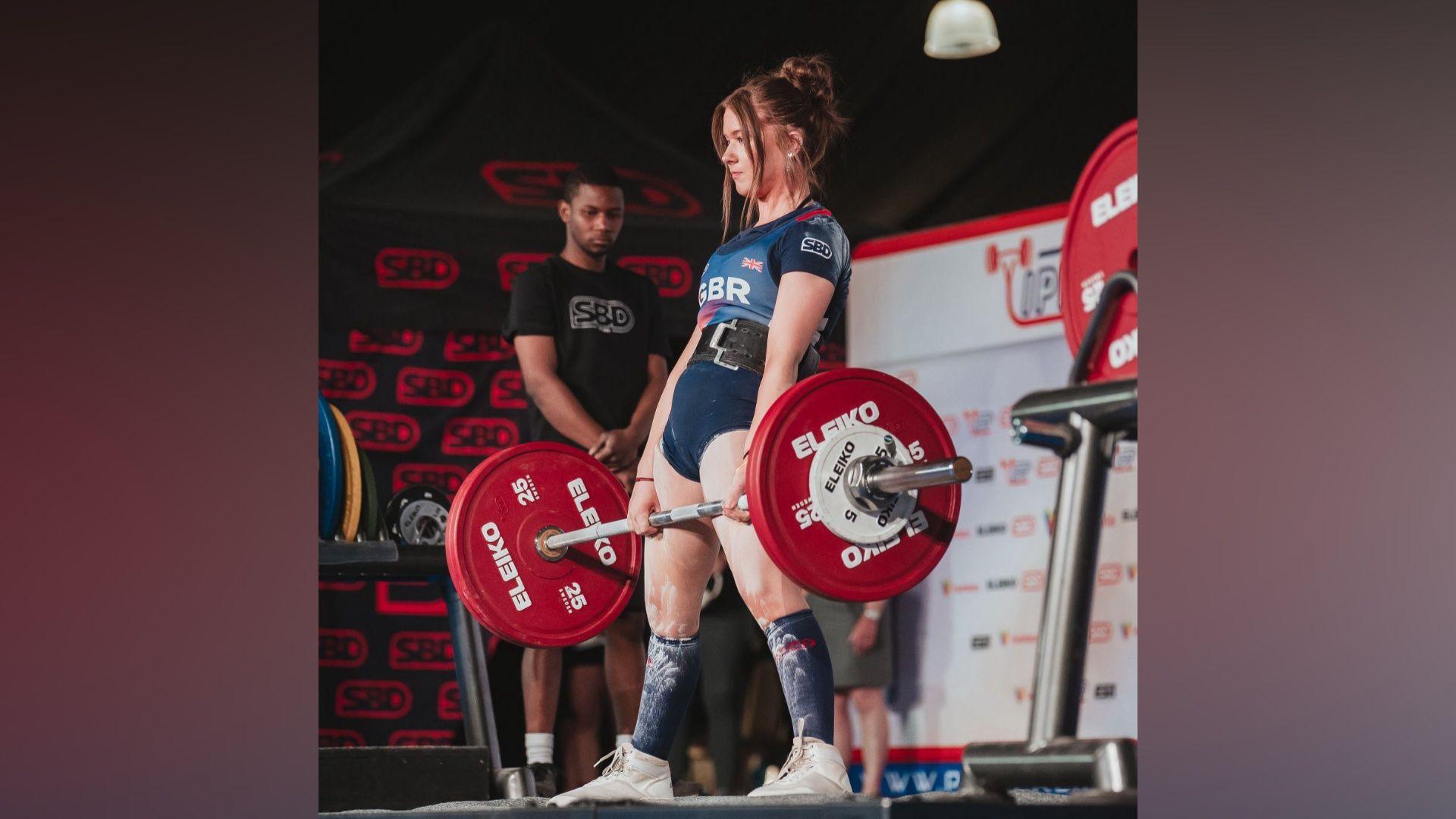 A female athlete dressed in a Great Britain tshirt, shorts, socks and trainers is lifting a weight bar whilst being overlooked by a competition umpire.
