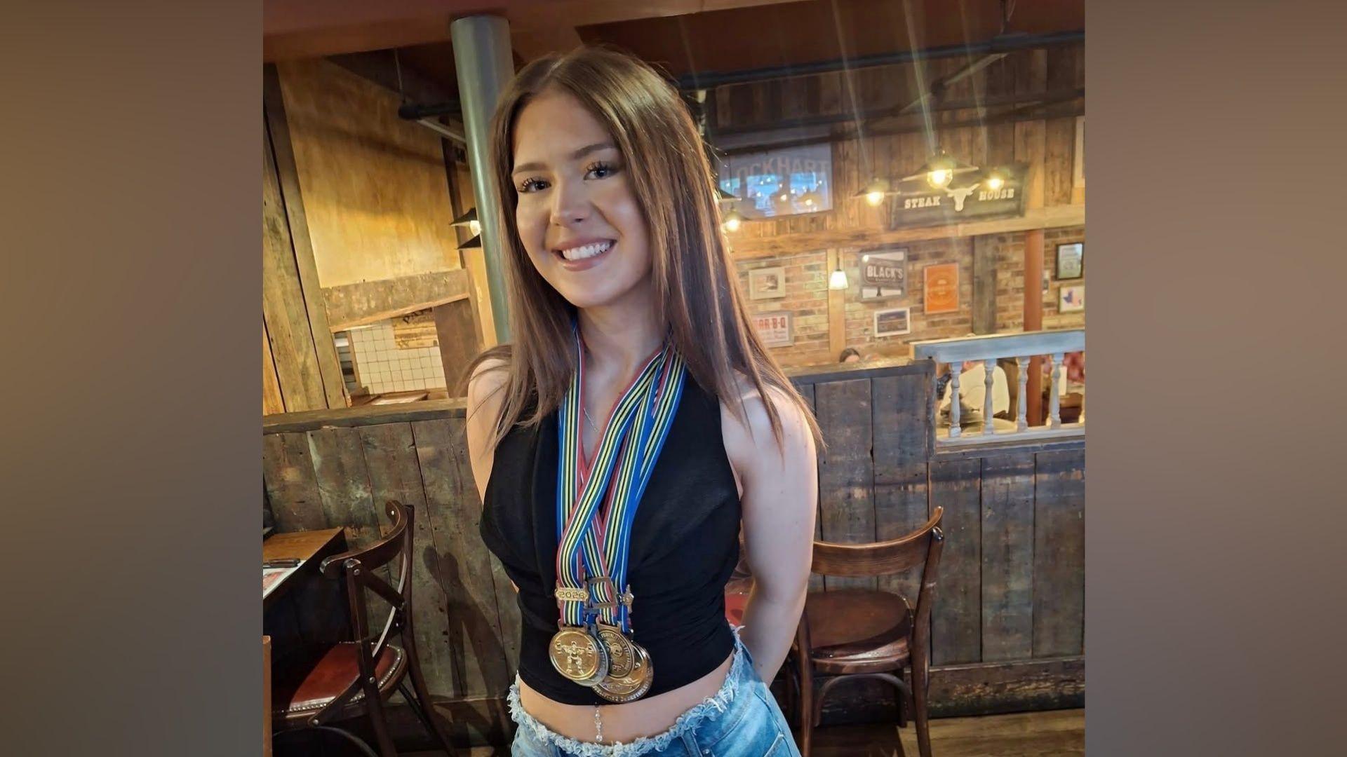 A young woman with long brown hair poses for a photo in a restaurant. She is wearing denim shorts and a black sleeveless top and has two gold and one bronze medals around her neck.