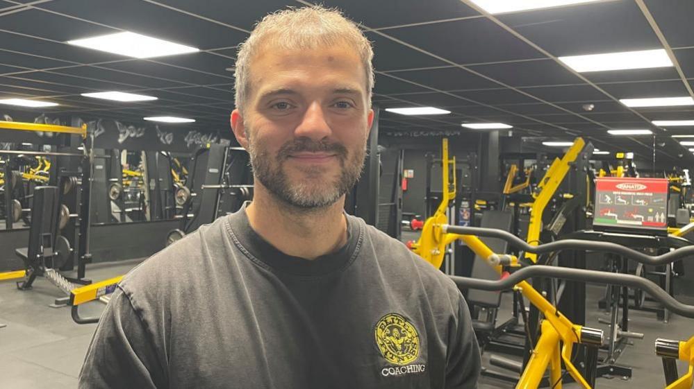 A man with short grey hair and beard smiles at the camera, wearing a grey t-shirt with a yellow logo for Elite Fitness Coaching. Behind him is a gym floor with multiple weight machines that are black and yellow.