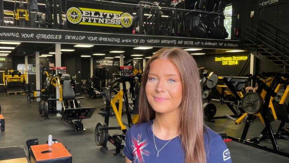A young women with long brown hair, wearing a blue top with a union jack flag on it smiles at the camera. Behind her is a gym across two floors with yellow and black weight machines.