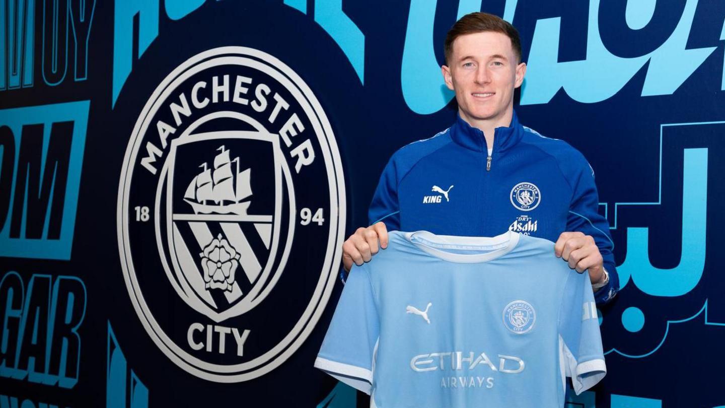 Manchester City midfielder Elliot Anderson poses with his new shirt infront of a club-branded wall