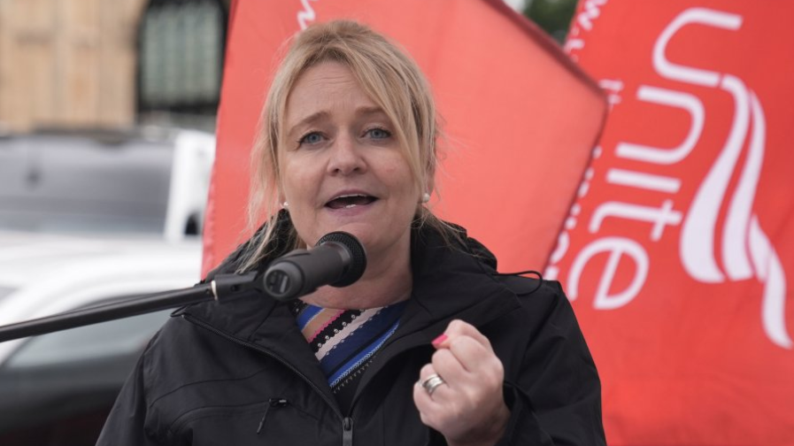 Head and shoulders shot of Graham, who has blonde hair pulled back in a ponytail and is wearing a black raincoat over a striped blue and black top. She has her fist clenched with red nail varnish visible and two wedding rings. She is standing in front of a red Unite the Union flag.