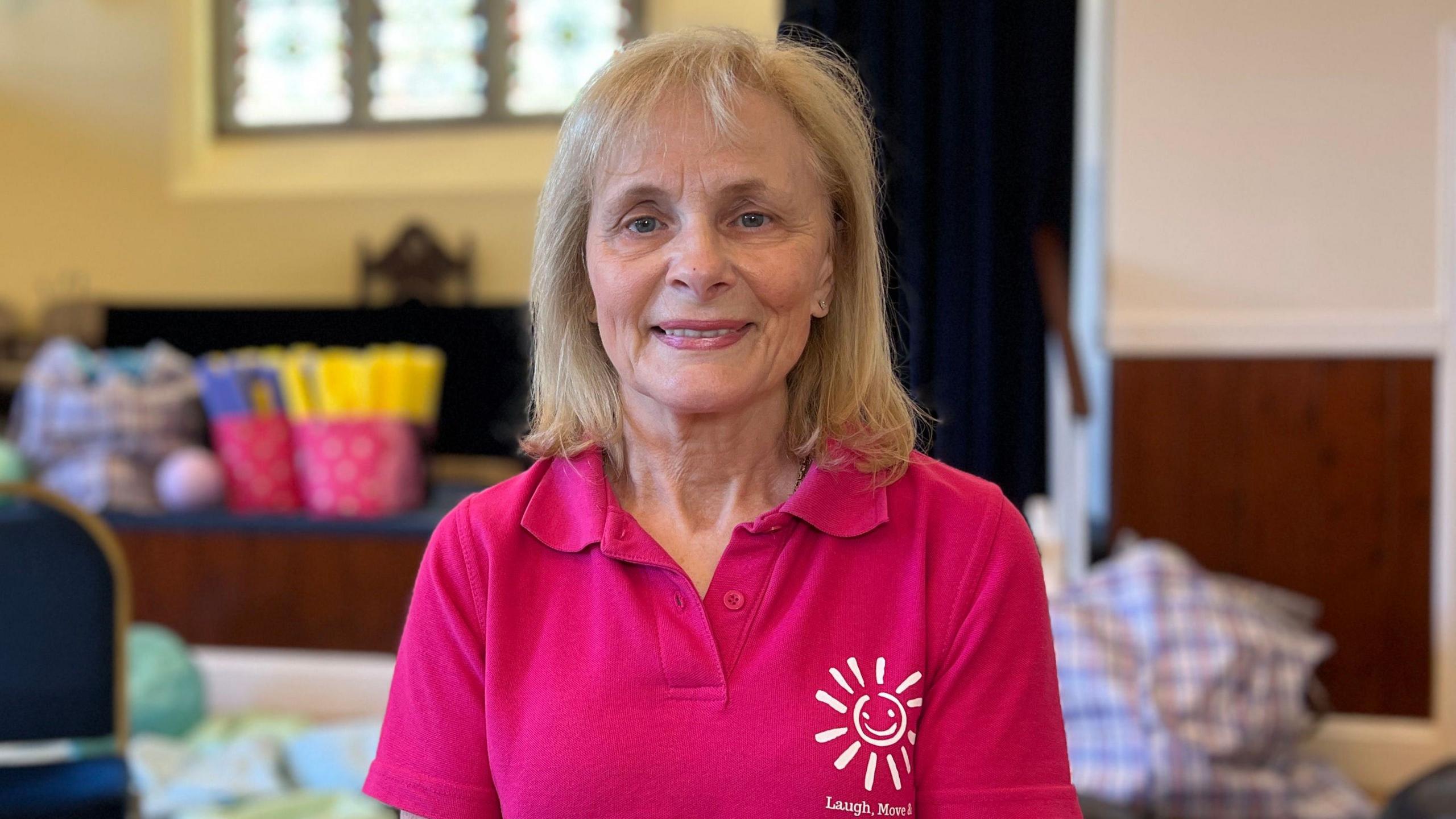 Jackie Turner is wearing a pink top and sits on a chair in a community hall, facing the camera. Empty chairs, radiators, large windows and wooden memorial plaques line the room behind her.