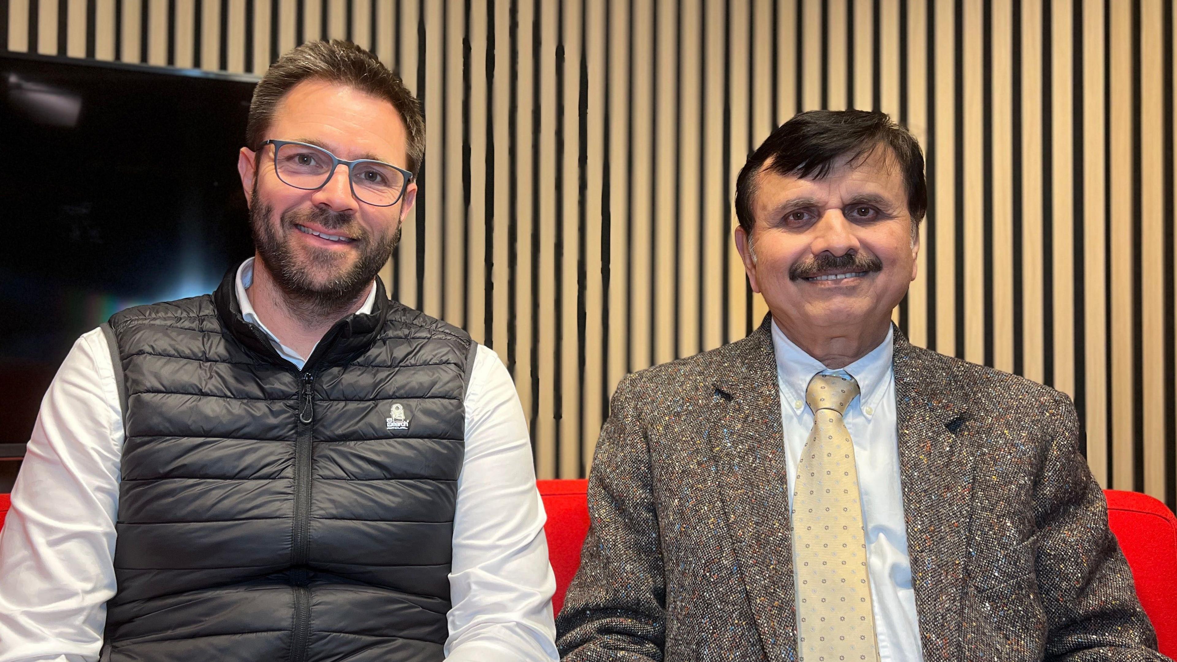 Marcus Liddiard and Dr Safraz Jamali sit side by side facing the camera.
Liddiard on the left is wearing a white shirt and a black gilet He has short brown hair and light-coloured spectacles. Jamali is on the right and wearing tweed-style jacket in mixed grey and brown tones, a light blue shirt and a light-coloured patterned tie. 