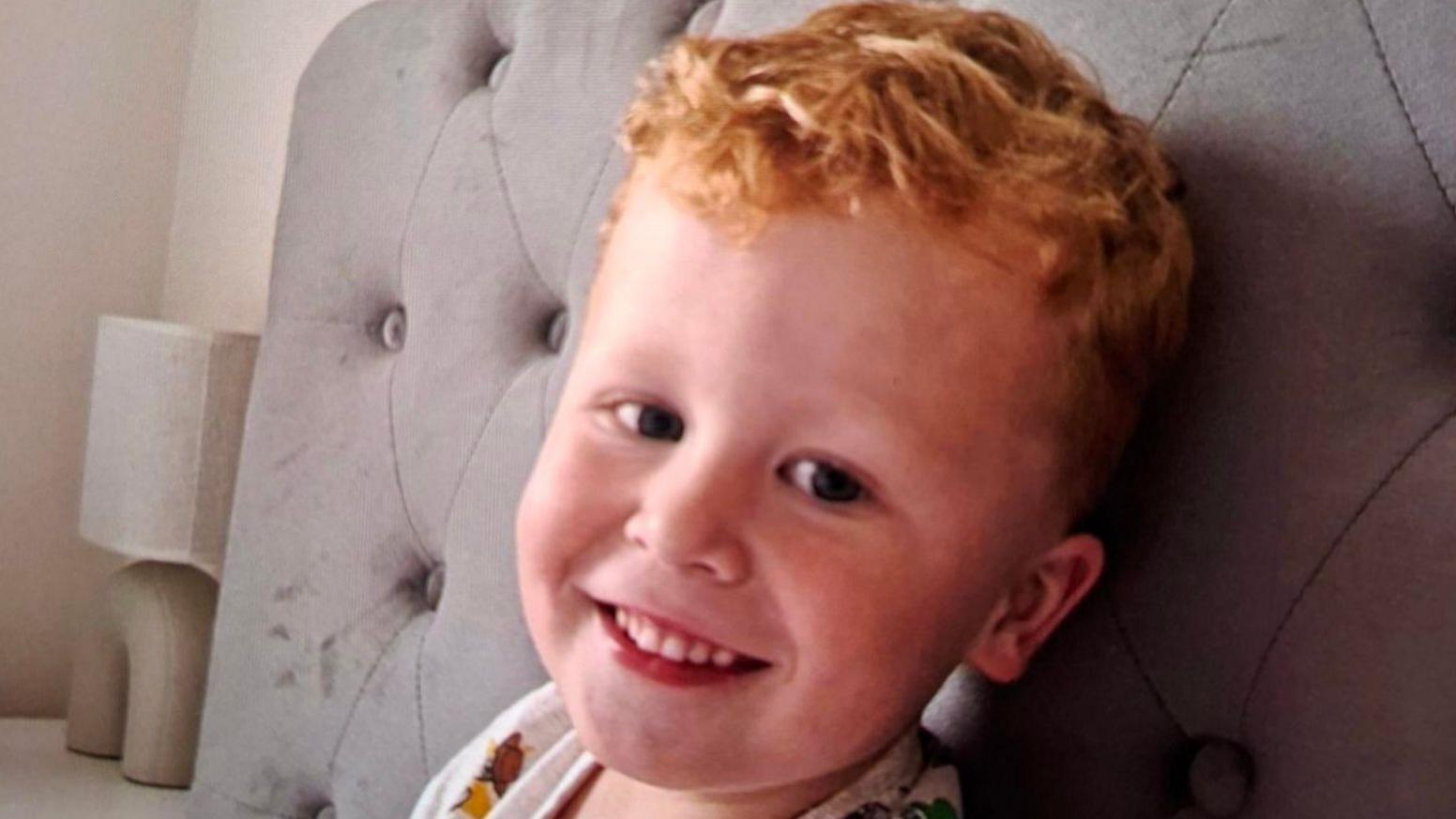 Three-year-old Noah Woods, with light hair and wearing a brightly coloured top, smiles at the camera.