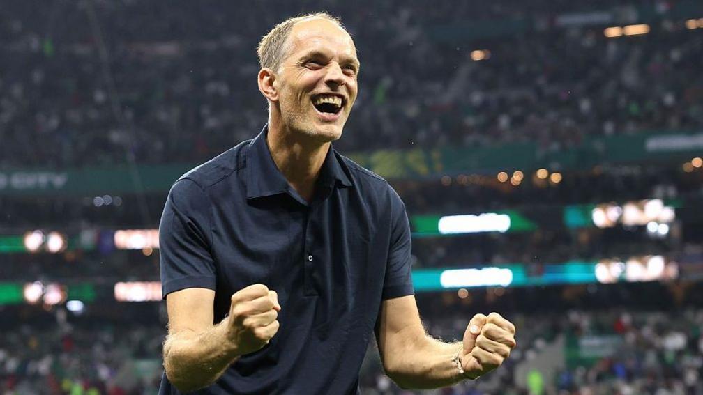 Thomas Tuchel, wearing a dark polo shirt, clenches two fists and grins in celebration