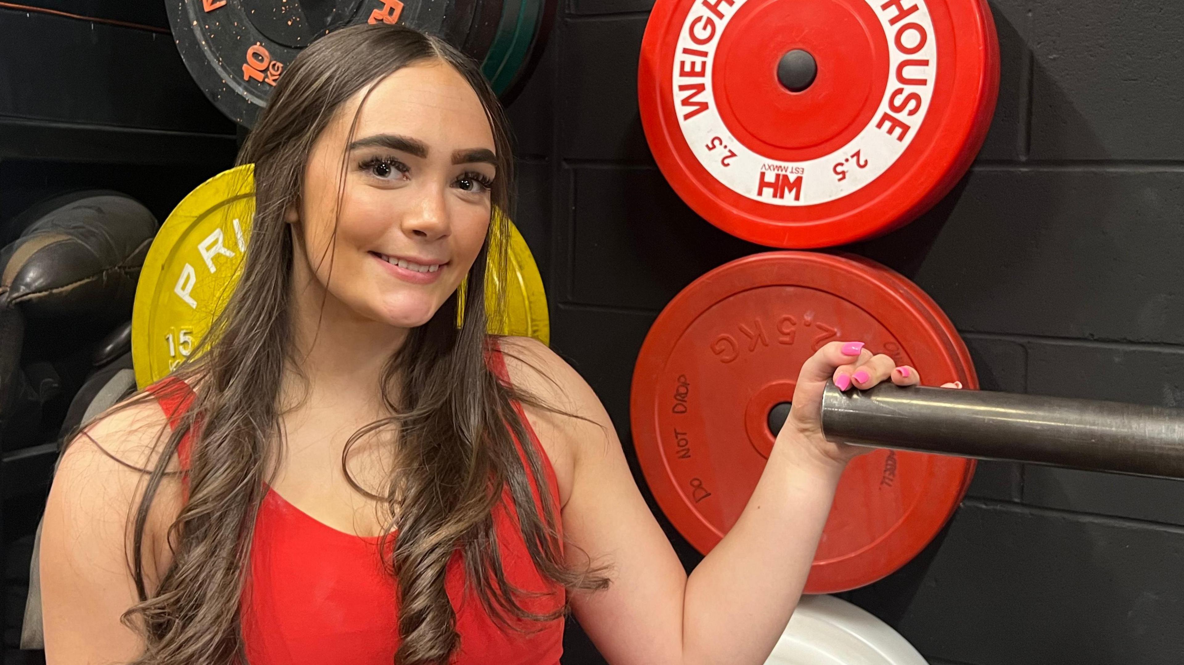 Annie, stood next to a weight rack wearing a red gym set. She has lots of different weights behind her as she smiles at the camera.