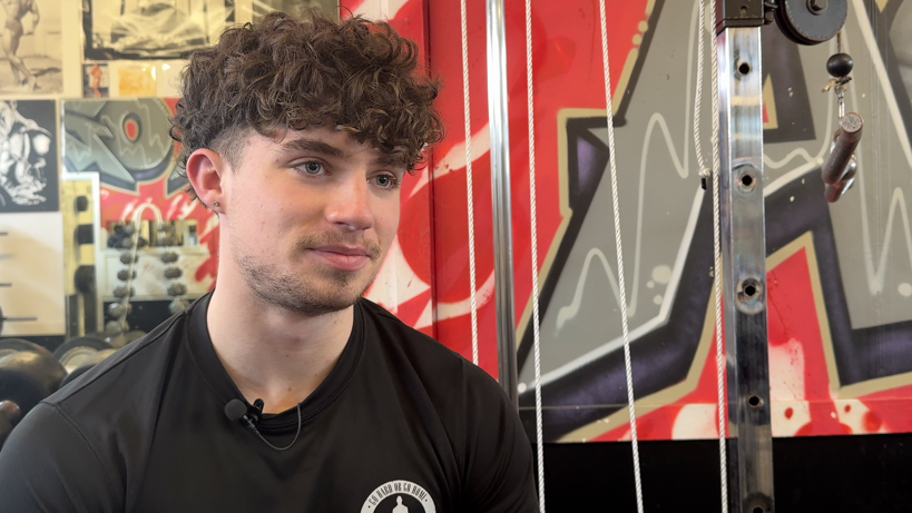 Charlie wearing a black t-shirt sat by gym equipment with graffiti on the wall behind