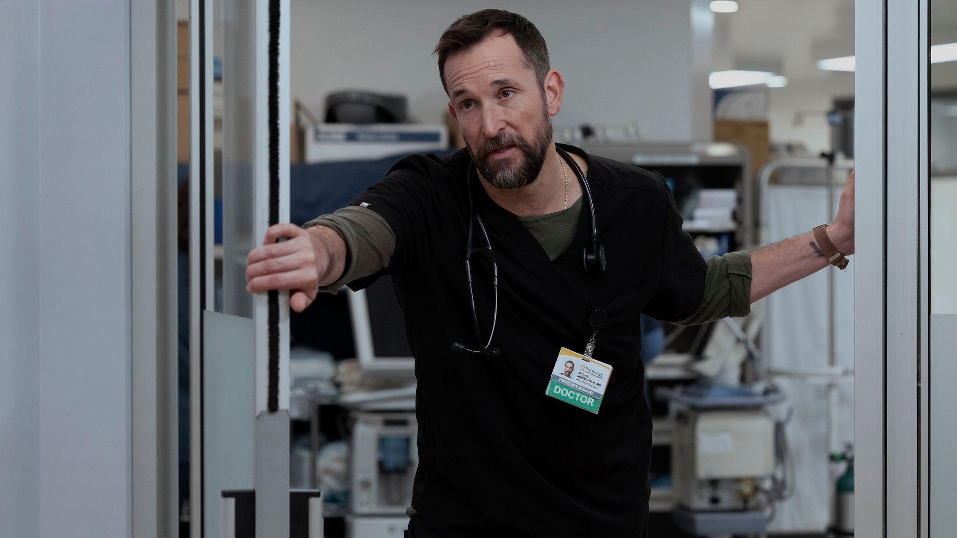 Dr. Michael “Robby” Robinavitch (Noah Wyle) looking exhausted, wearing black scrubs and holding a door open