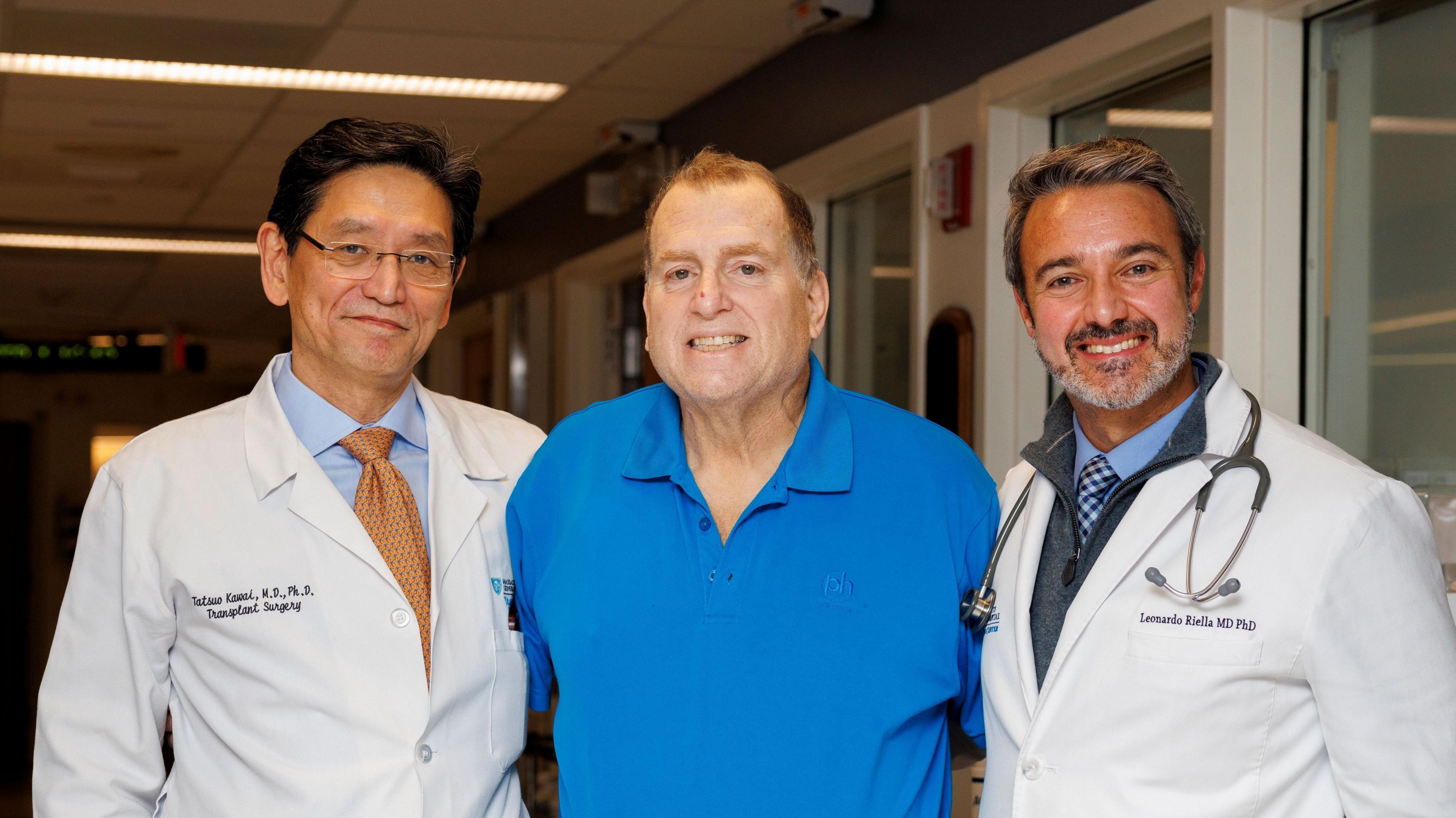 The image shows three adults standing closely together in a hospital hallway. The person in the centre is wearing a bright blue polo shirt, while the individuals on the left and right are wearing white medical coats, with a stethoscope visible around the neck of the person on the right. Fluorescent ceiling lights illuminate the corridor, and doors and windows line the walls behind the group. Small embroidered text is visible on the medical coats.