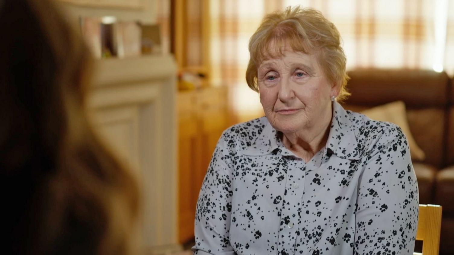 A woman with fair hair and wearing a grey and black patterned top looks at a person off camera. She is sitting in a living room with curtains drawn.