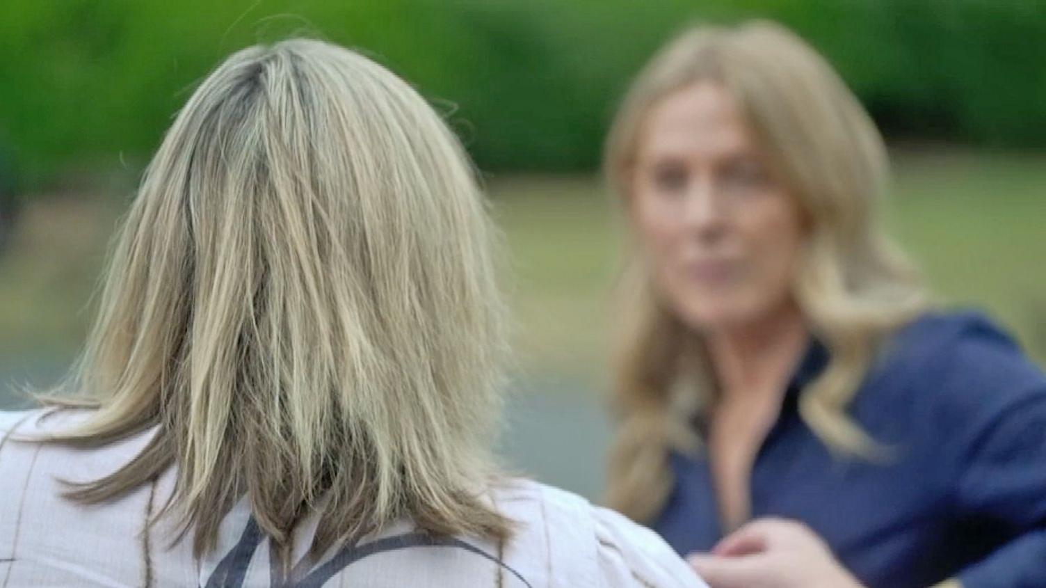 A woman with fair hair - photographed from behind - looks at another woman who is out of focus.