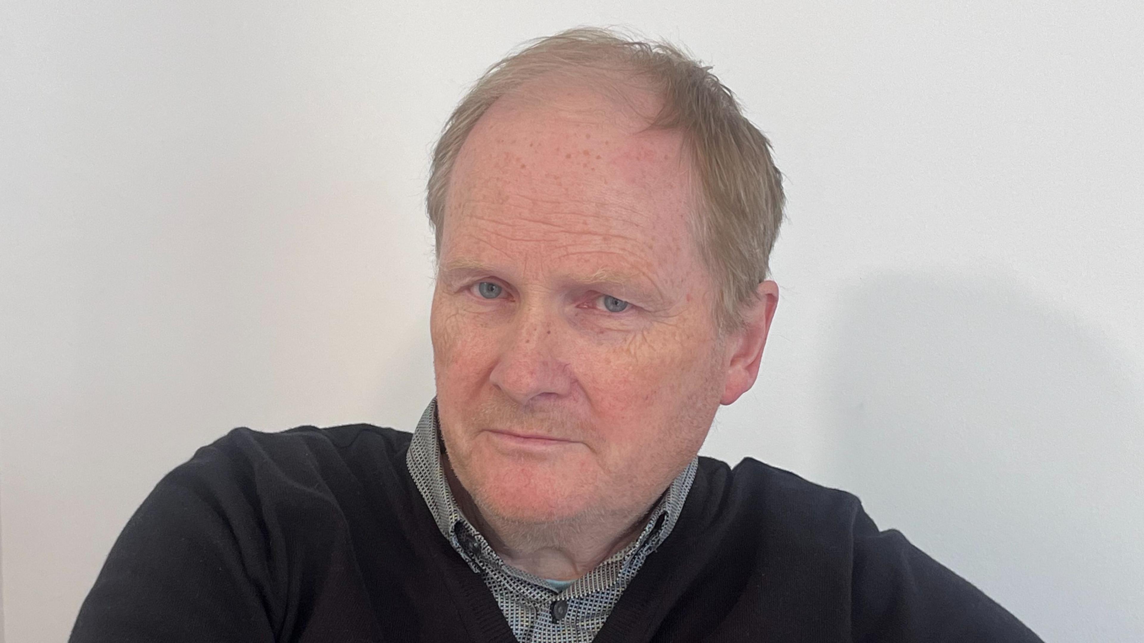 William Duncan is an older man with short fair hair that is thinning on top of his head. He is wearing a grey shirt with a black v-neck jumper over it and is sitting in front of a plain white wall. 