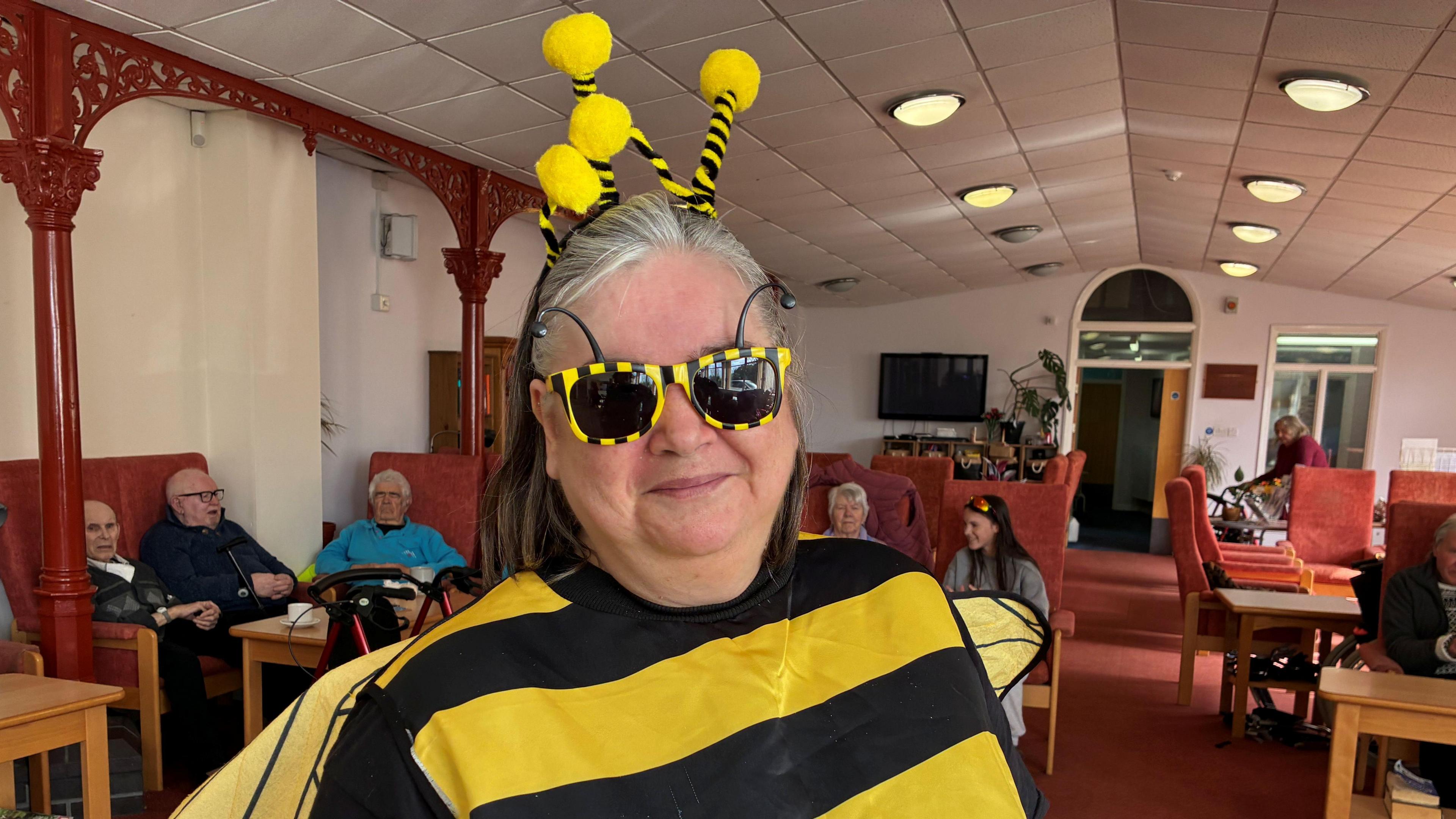 A woman in a fancy dress bee outfit and black and yellow sunglasses smiles at the camera, behind her are a group of elderly people sitting in chairs at a day centre. 