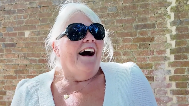 A woman with blonde hair and big black sunglasses smiles while speaking to the camera