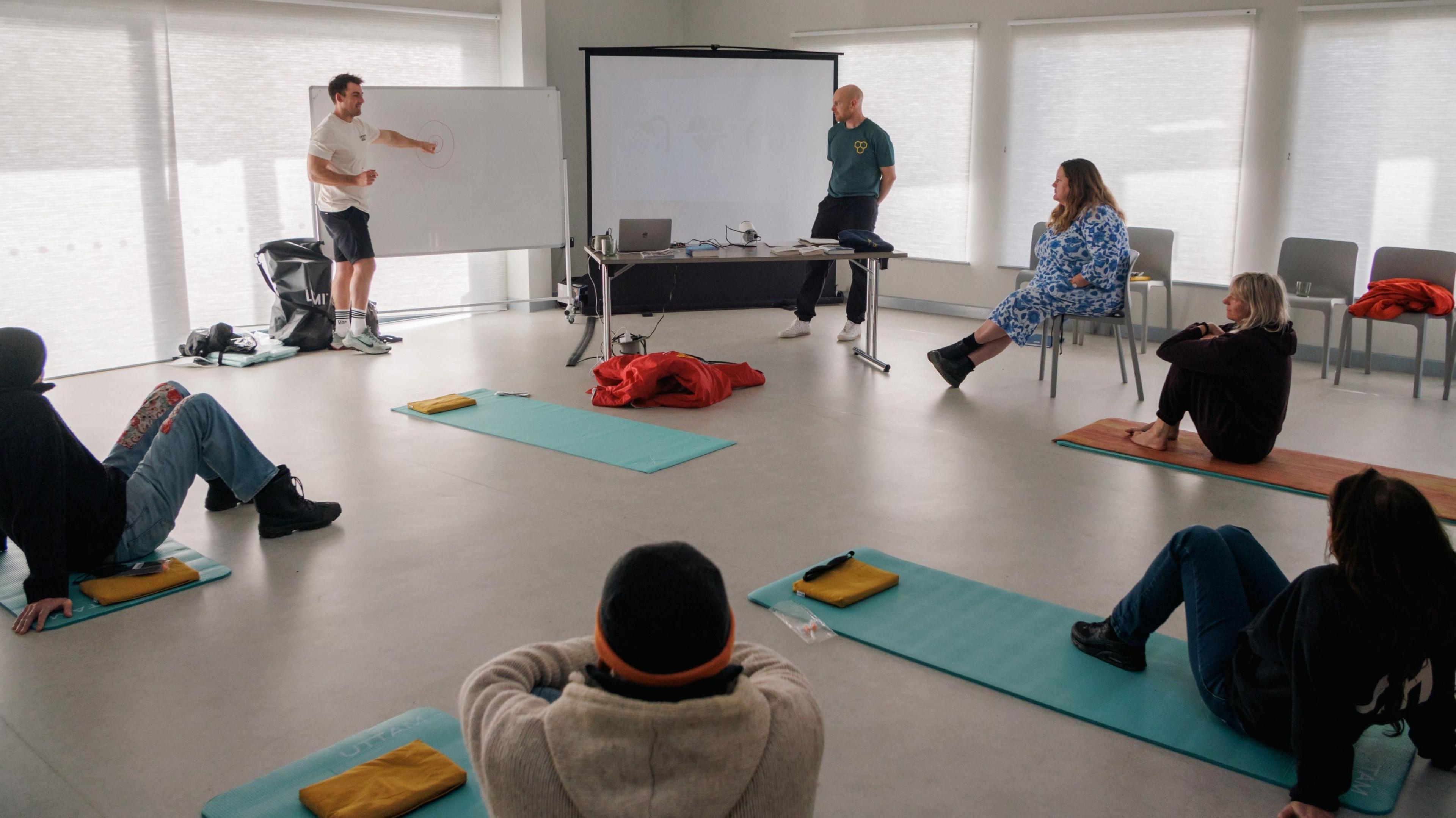 A room with a grey hard floor and white blinds covering the windows on the edges of the room. There are 5 turquoise-blue yoga mats on the floor with people sat on four of them, and two instructors stood near a board presenting a class of sorts. There is also a woman in a blue dress sat on a chair.