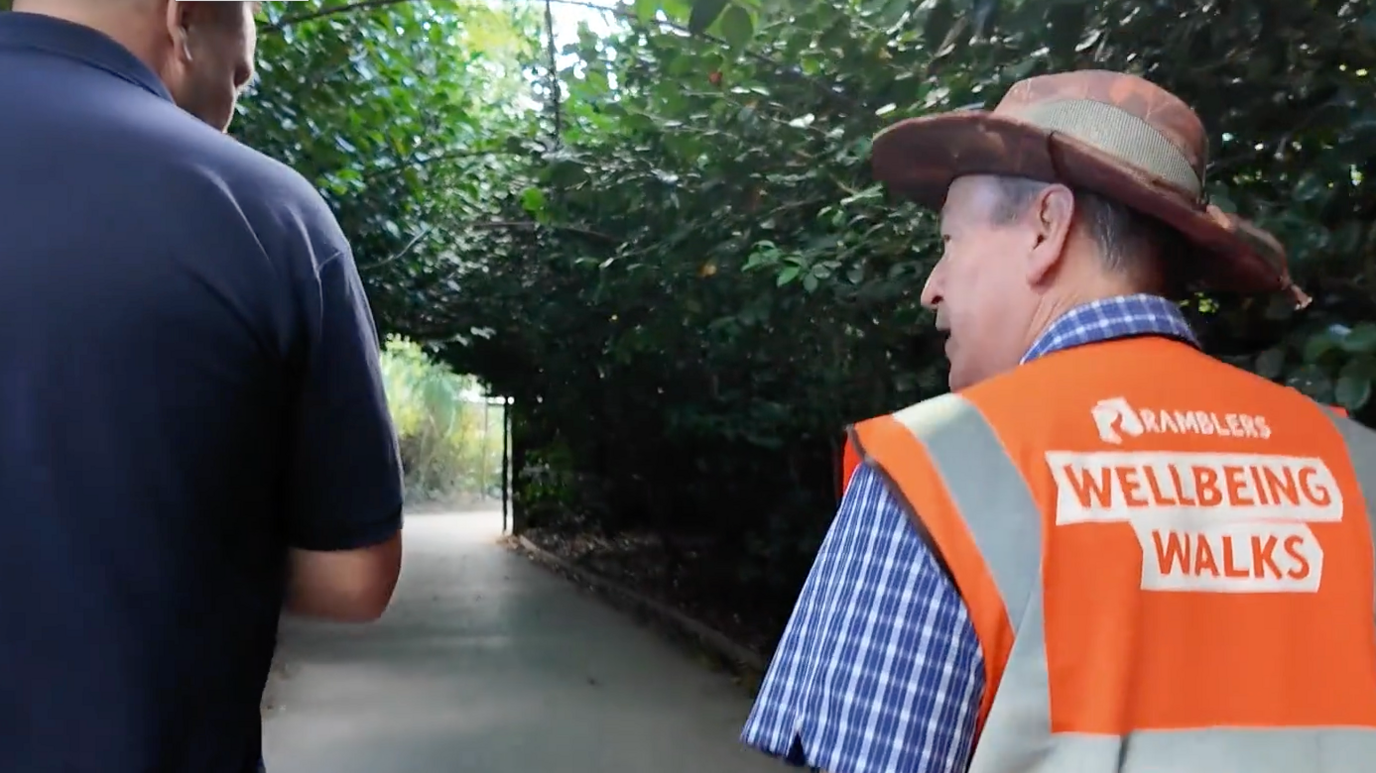 A man in an orange high vis jacket which reads 'Ramblers Wellbeing Walks' chats to another man in a navy polo shirt while walking. They are walking with their backs to the camera.