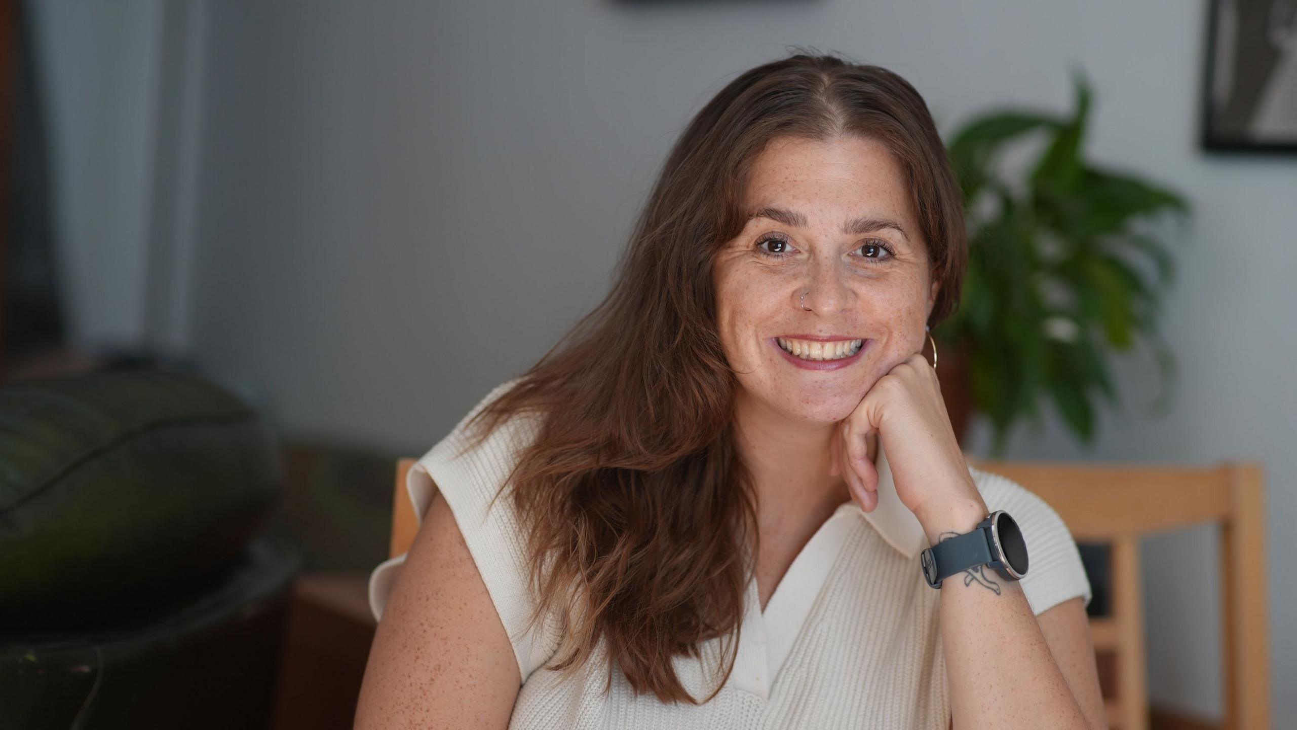 Jessica Hill, a woman in her 30s with long red-brown hair, smiles at the camera while resting her chin on her hand; she's wearing a white short-sleeved shirt with a collar, and a Garmin smartwatch on her wrist. She's sitting on a wooden chair, and a large house plant can be seen in the background, out of focus.