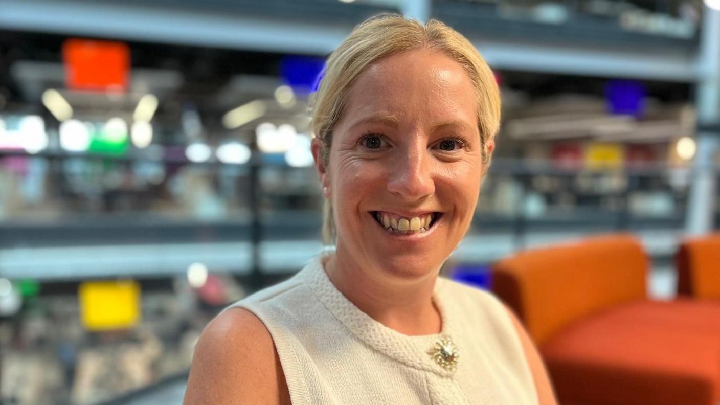 Michelle is smiling at the camera, she is wearing a cream sleeveless top and is sat on orange seats, with an open-plan, multi-level office setting in the background.