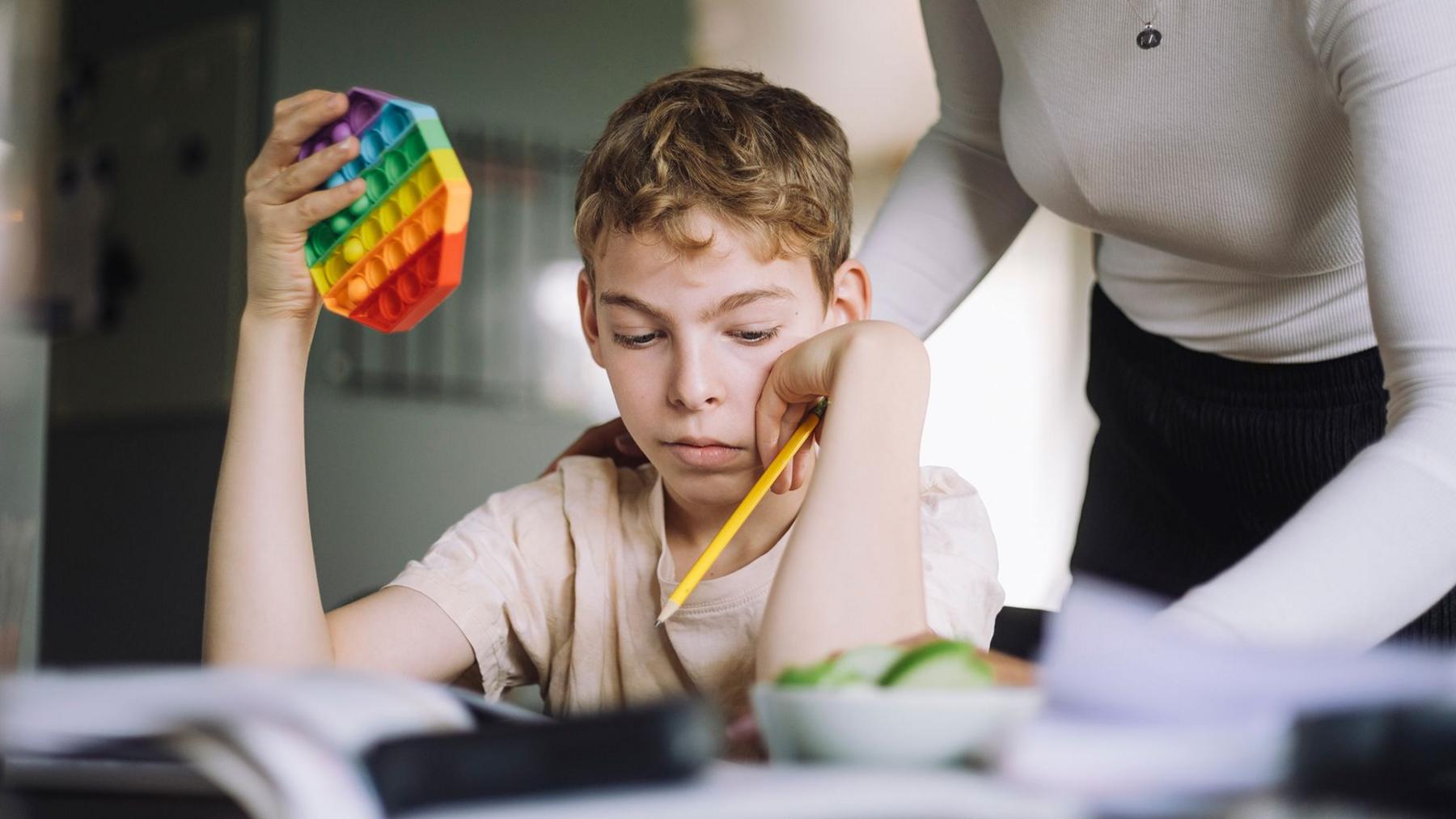 Young boy sits at table with pencil in one hand and colourful puzzle in the other. A woman stands over him.