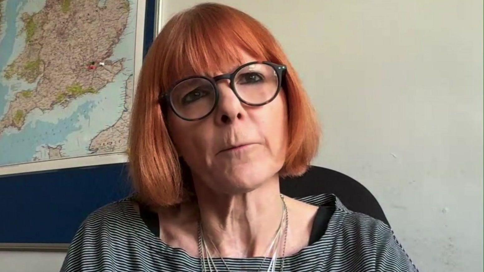 Jane Fisher has shoulder length red hair. She is sitting on a chair in front of a white wall and a map of the UK. She is wearing round black glasses with a necklace and a white and black striped top. 